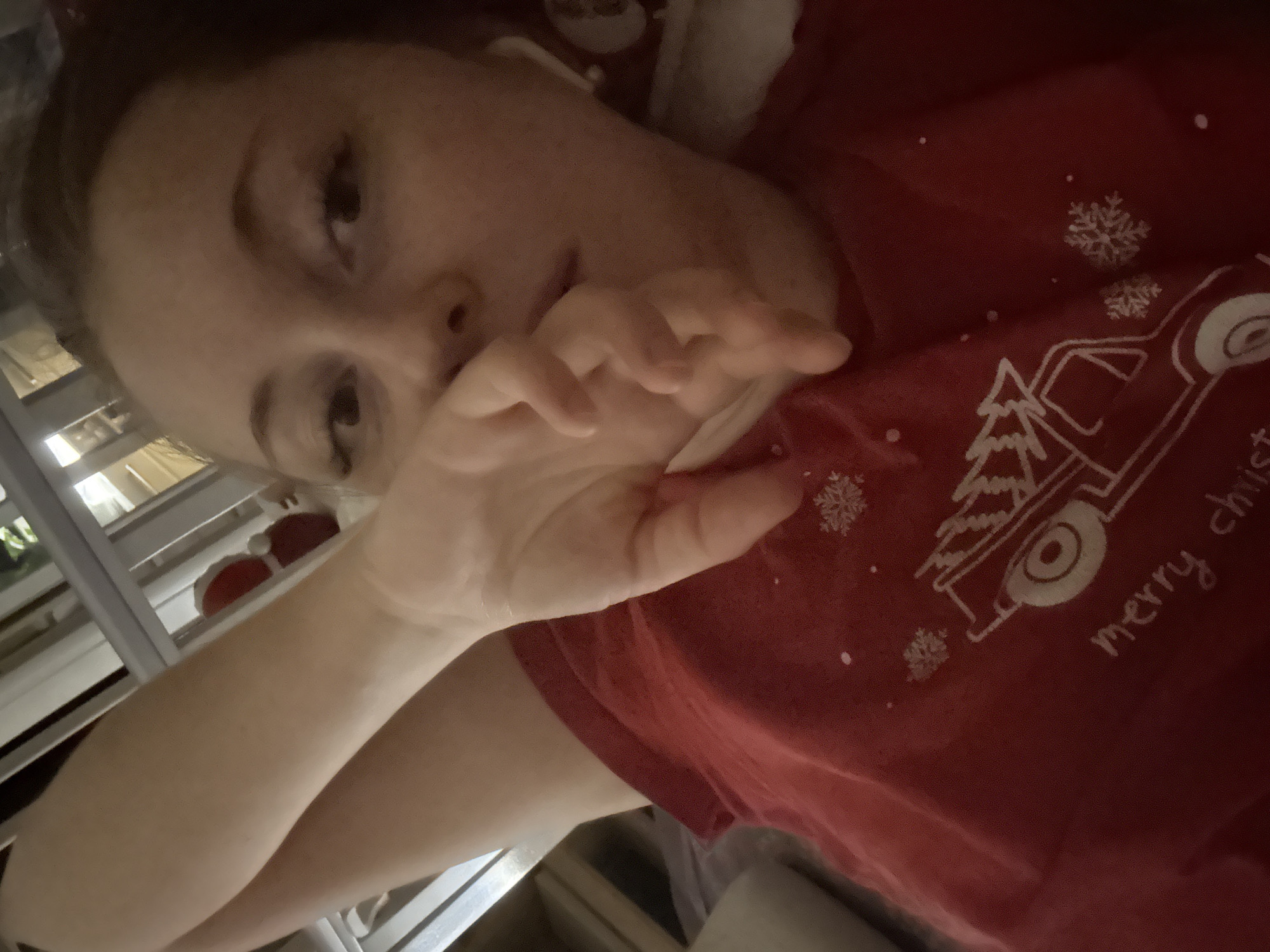 An overhead close-up selfie features a person, likely female, lying down indoors. She is wearing a red t-shirt adorned with white outline designs, including a vintage-style truck carrying a Christmas tree, several snowflakes, and the partial text "merry christ" (likely "merry christmas"). Her left hand is casually positioned near her chin or mouth, partially obscuring her lower face. Her face is tilted, and one eye is clearly visible, looking directly towards the camera, conveying a relaxed and candid expression. The lighting is soft and dim, suggesting an indoor setting during evening or low-light conditions. The background is out of focus, showing indistinct vertical and horizontal elements, possibly parts of a window frame or furniture, indicating an interior space.