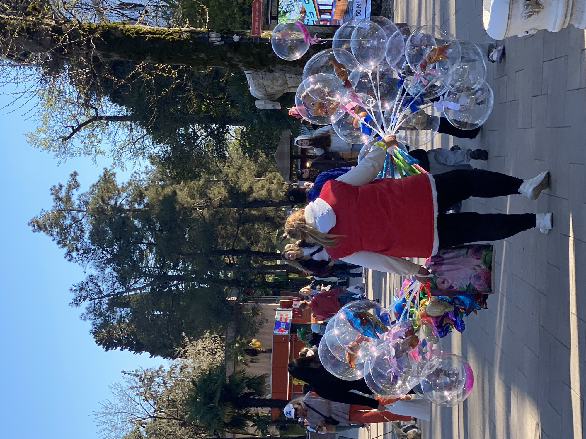 A street scene shows a woman standing sideways, holding a large bunch of clear balloons with various small objects inside. She is wearing a red vest over a white long-sleeved shirt, black pants, and white shoes. The background includes a sidewalk, trees, and other people. A building is visible in the distance. The location is likely Sochi, Russia.