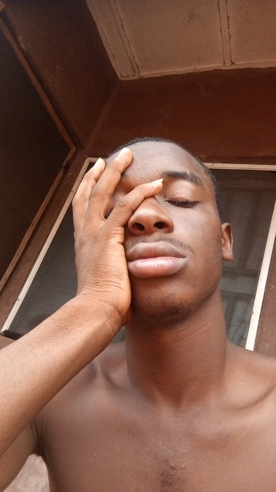 A close-up portrait features a young man with dark skin, likely of African descent, captured from the chest up. His eyes are closed, and his right hand covers a portion of his face, specifically his right eye and forehead, conveying an expression of weariness, contemplation, or perhaps a moment of emotional processing. His lips are full and slightly parted. He is bare-chested, revealing his shoulders and upper chest.
The lighting is natural and soft, suggesting it is daytime, illuminating his features and casting subtle shadows. The background is out of focus, showing dark brown and lighter beige elements that appear to be part of an interior or semi-outdoor structure, possibly a room or verandah. A window frame is partially visible behind him on both sides, suggesting proximity to a window. No discernible text is present in the image. The scene depicts a personal and quiet moment, potentially in a domestic setting in Onitsha, Nigeria.