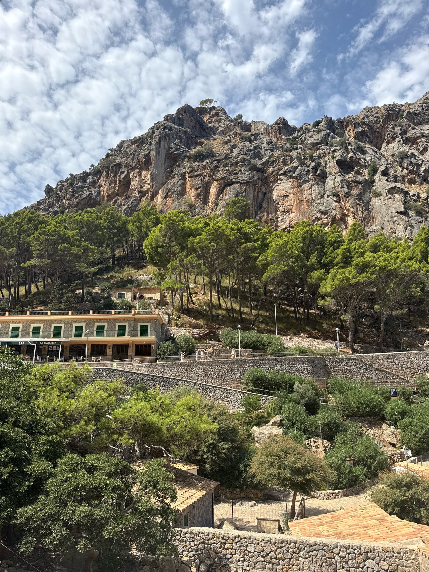 The image depicts a mountain landscape with a building and surrounding vegetation in Port De Sa Calobra, Spain. In the foreground, a stone wall borders a pathway, with several terracotta-tiled roofs visible among trees and shrubs. A two-story building, likely a restaurant or hotel, is situated on a terrace, characterized by its stone facade, green shutters, and an awning that reads "LA CALOBRA BAR". Several tables and chairs are arranged on the patio in front of the building. A steep, rocky mountain slope covered with pine trees rises behind the building. The sky is partly cloudy with a mix of white and grey clouds against a blue background. Two people are visible walking on a path near the building, and another person is walking on a lower path.