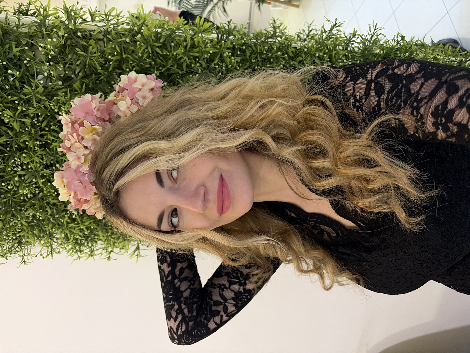 A young woman with blonde, wavy hair and a flower crown poses indoors against a backdrop of artificial greenery. She wears a black lace top or dress. The setting appears to be a brightly lit room. There is no specific evidence within the image to confirm the location is in Budva, Montenegro.