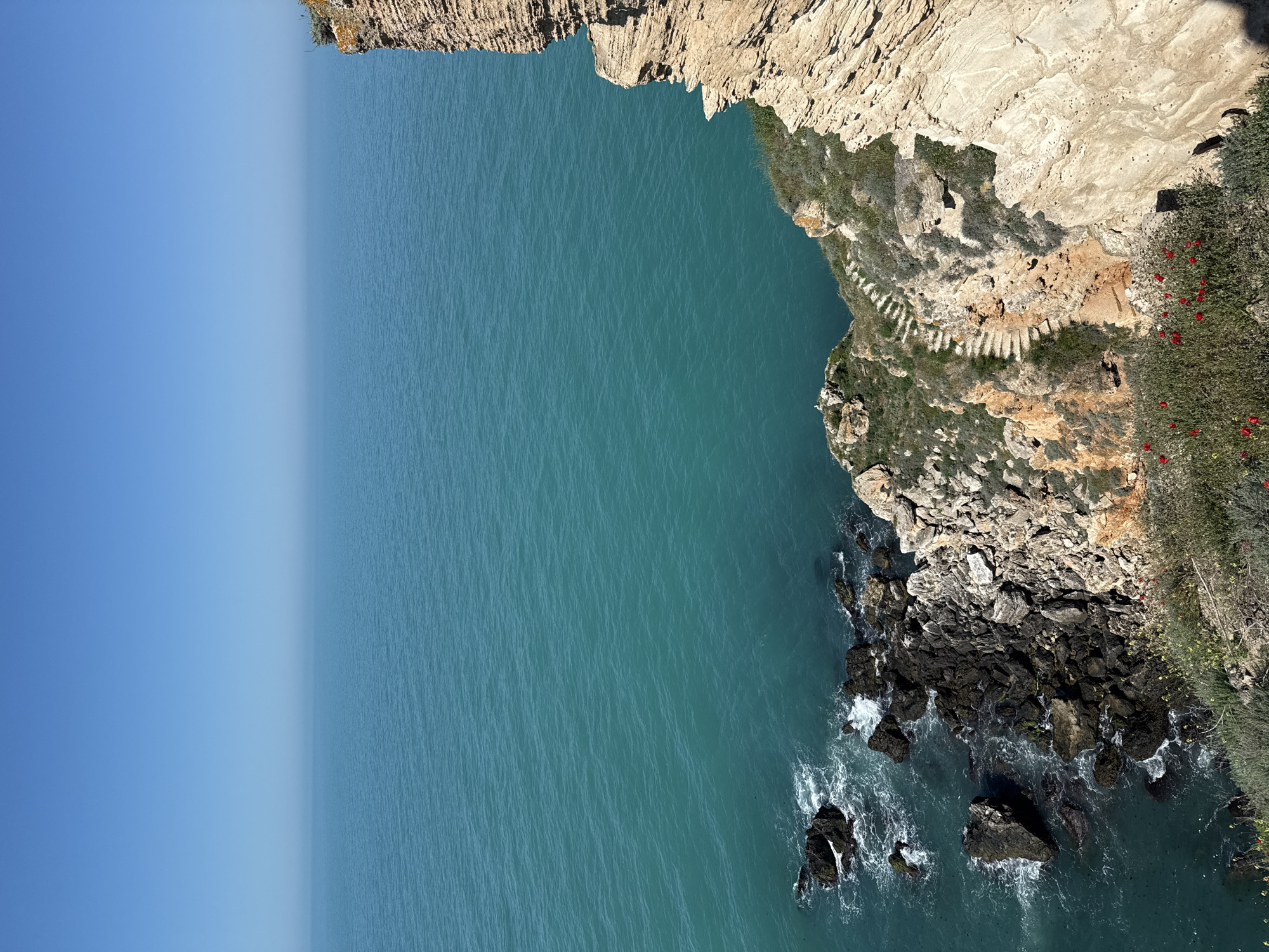 The image shows an aerial view of a rocky coastline in Balgarevo, Bulgaria. The scene includes a steep, light-colored cliff face with vegetation growing along its edge. A set of stairs is carved into the cliffside. The turquoise water meets the rocky shore, and waves crash against the dark-colored rocks at the base of the cliff. The sky is blue.