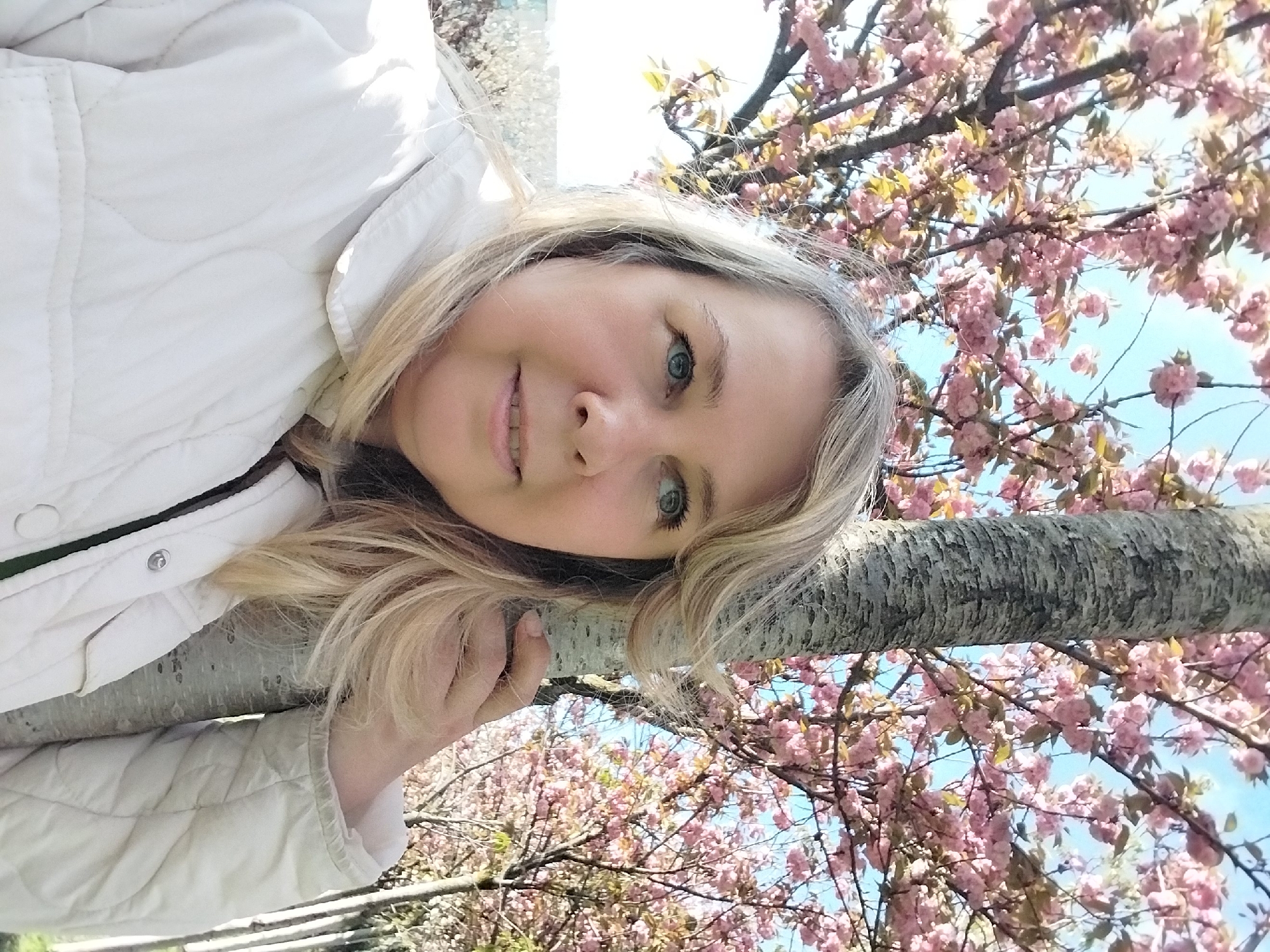 A rotated close-up portrait shows a blonde woman with blue eyes, smiling softly at the camera. She is wearing a white quilted jacket. Her head is tilted, and her right hand rests gently on a thick tree trunk. The background is filled with abundant pink blossoms on tree branches, set against a clear blue sky, indicating a bright, sunny spring day. The overall impression is one of natural beauty and contentment. The scene is outdoors, likely in a park or garden in Sochi, Russia. There is no visible text.