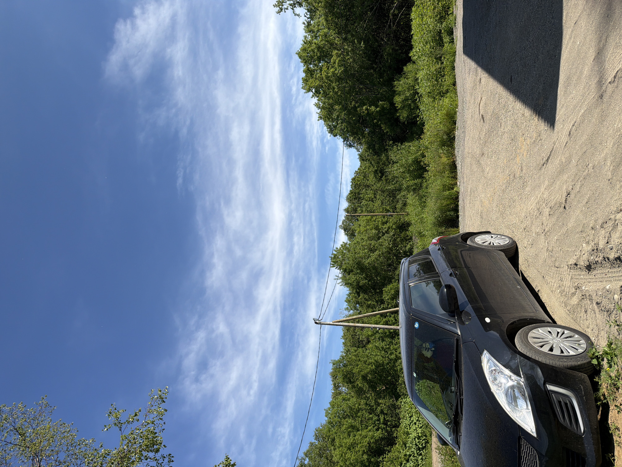 A black hatchback car is parked on a dirt lot in the city of Novy, Russia, with a dense line of green trees and foliage forming the background. The sky above is a vibrant blue, dotted with thin, wispy white clouds. A wooden utility pole with wires is visible in the middle ground, slightly to the left of the car. The car's front left side is angled towards the viewer, showcasing its headlights, grille, and front wheel. The dirt lot has tire tracks imprinted on its surface. A dark shadow from an unseen structure falls across the right side of the frame.
