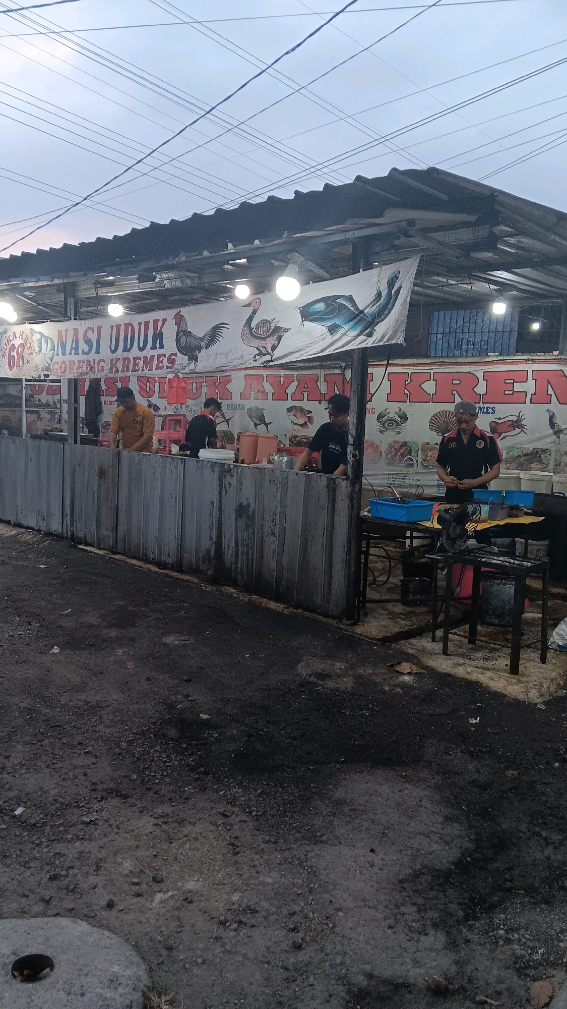 The image depicts an outdoor food stall in Depok, Indonesia, likely operating in the evening or early morning given the overcast sky and artificial lighting. The stall features a long, gray metal counter that separates the serving area from the customer's space. Behind the counter, several people are visible working. One person in a yellow shirt is preparing food, while another in a black shirt appears to be serving. Further to the right, a man in a black uniform with a gray cap is standing near a table with cooking equipment.
A large banner hangs above the stall, advertising "NASI UDUK GORENG KREMES" and "NASI ULUK AYAM KREMES," indicating that fried rice and crispy fried chicken are the main offerings. The banner also features illustrations of poultry and fish. The stall is situated on a dirt and gravel surface, with evidence of charcoal and ash on the ground, suggesting active cooking. The environment appears somewhat rustic, with electrical wires strung across the top of the frame and a corrugated metal roof covering the stall. The overall impression is of a busy, local eatery.