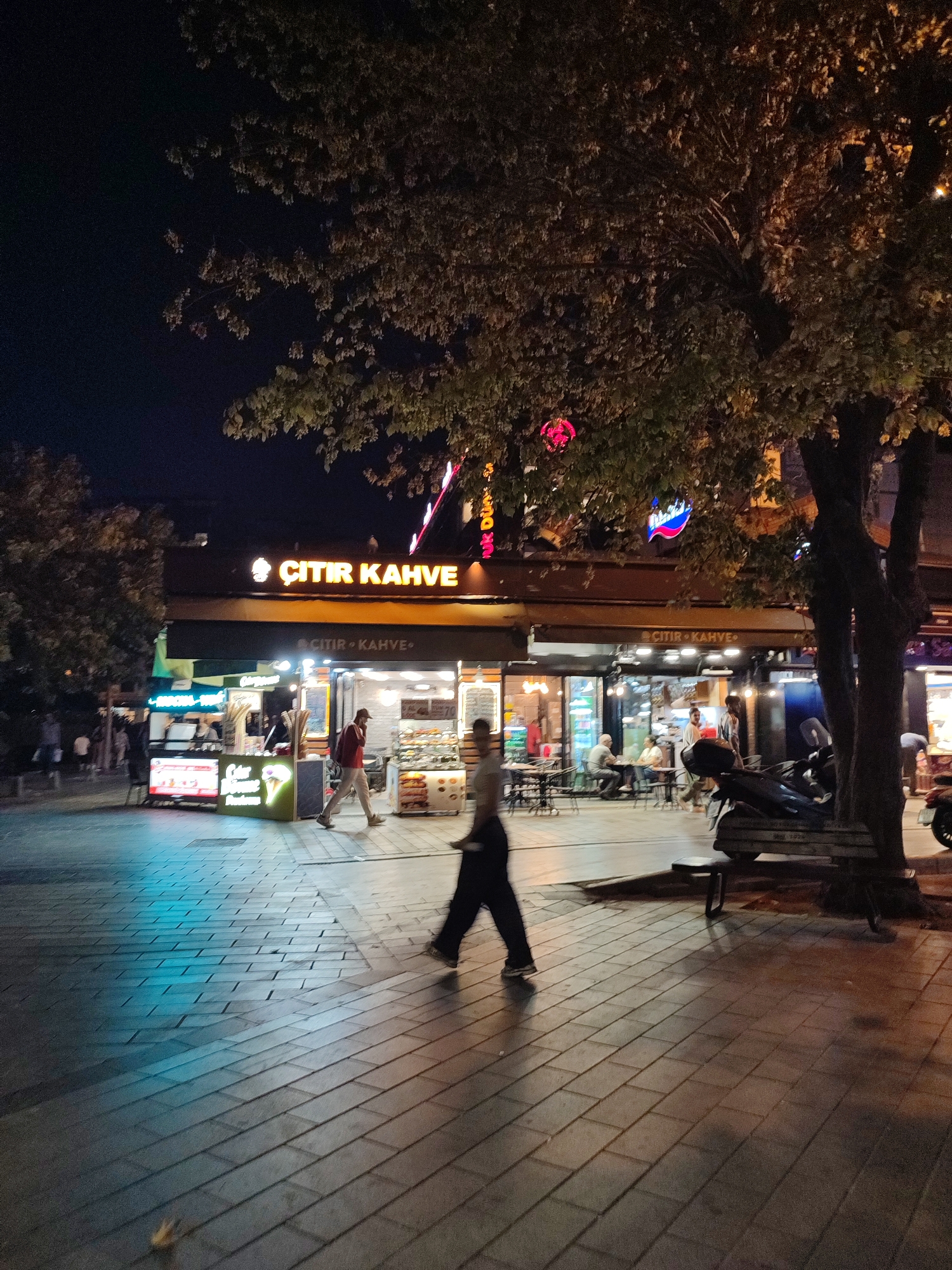 The image depicts a nighttime street scene in Üsküdar, Turkey, focusing on a brightly lit cafe called "ÇITIR KAHVE." Several people are visible in the foreground and background. In the immediate foreground, a person is walking across a tiled sidewalk, their form blurred due to motion. To the left, a person in a red jacket and a light-colored hat is walking away from the viewer. Further back, individuals are seated at tables outside the cafe, engaged in conversation. The cafe's facade features illuminated signage and display cases showcasing food items, likely pastries and ice cream. A motorcycle is parked to the right of the cafe, beside a wooden bench. The street is paved with patterned tiles, and overhead, the branches of a large tree partially obscure the night sky and decorative lights. The overall atmosphere suggests a lively evening in a commercial district.
