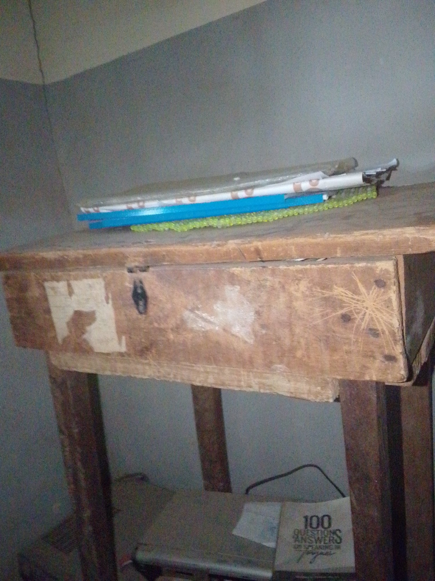 The image shows a wooden desk with a single drawer in what appears to be a room. On top of the desk, there is a stack of flat items, including what looks like a blue ruler and a green beaded item. The drawer of the desk has some wear and tear, with visible scratches and discoloration on its surface. There is also a small black latch or lock on the drawer. Below the desk, a book is visible, with the text "100 QUESTIONS ANSWERS" and some other unreadable text on its cover. The walls of the room are painted a muted gray color. The overall impression is of a simple, utilitarian desk in a modest setting. The image does not provide specific details that would allow for precise localization within Jalingo, Nigeria, beyond the general context of a room in that city.