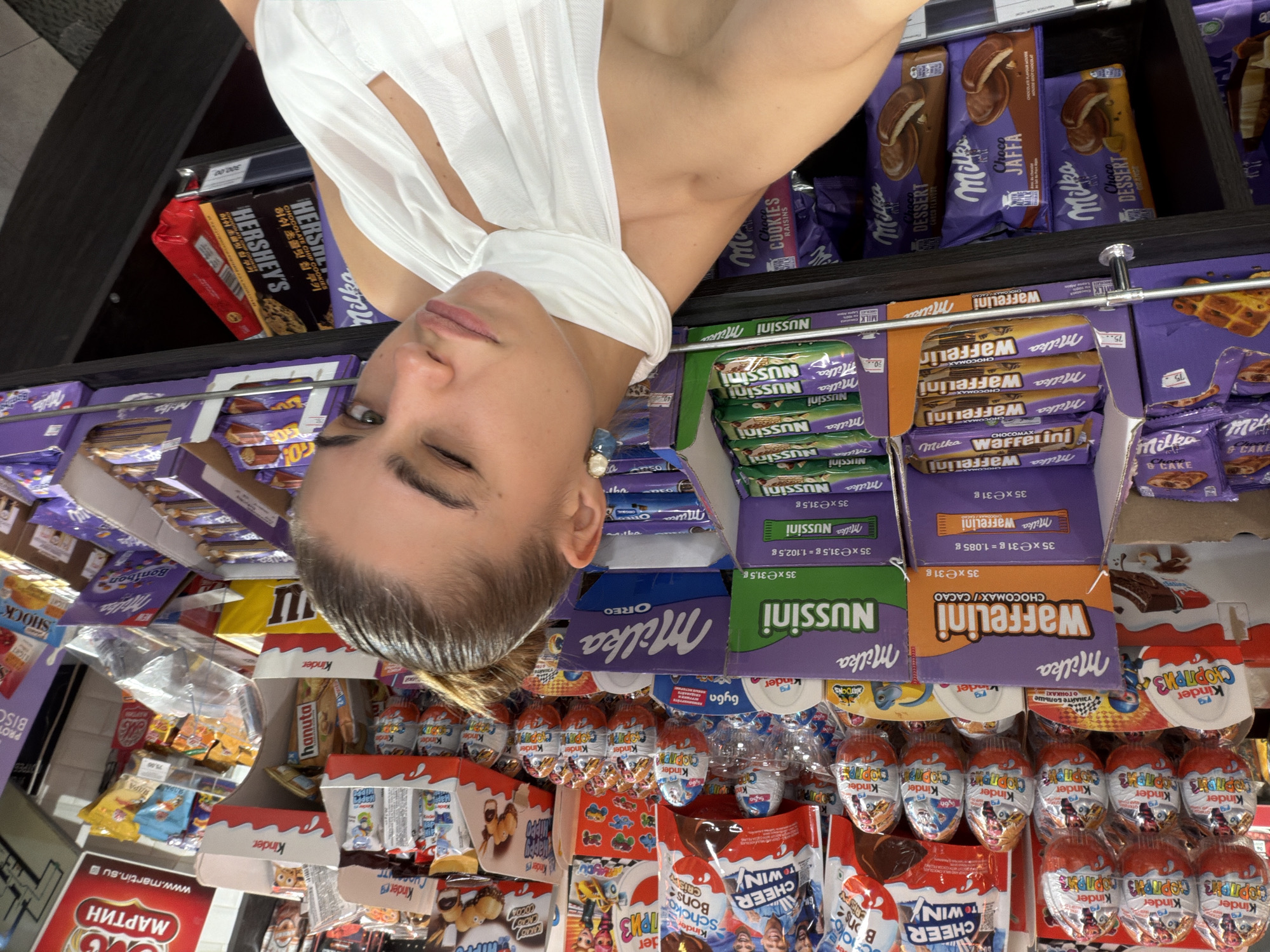 The image shows a young woman in a white top in front of a shelf filled with various chocolate products. The shelf displays brands such as Kinder, Milka Waffelini, Milka Nussini, Milka Oreo, and Hershey's. The products are packaged in colorful wrappers, predominantly purple and orange, and include items like chocolate eggs, bars, and cookies. The woman is looking towards the camera and winking. The environment appears to be a store aisle. The text on some of the products and signs indicates that the location is Moscow, Russia.
