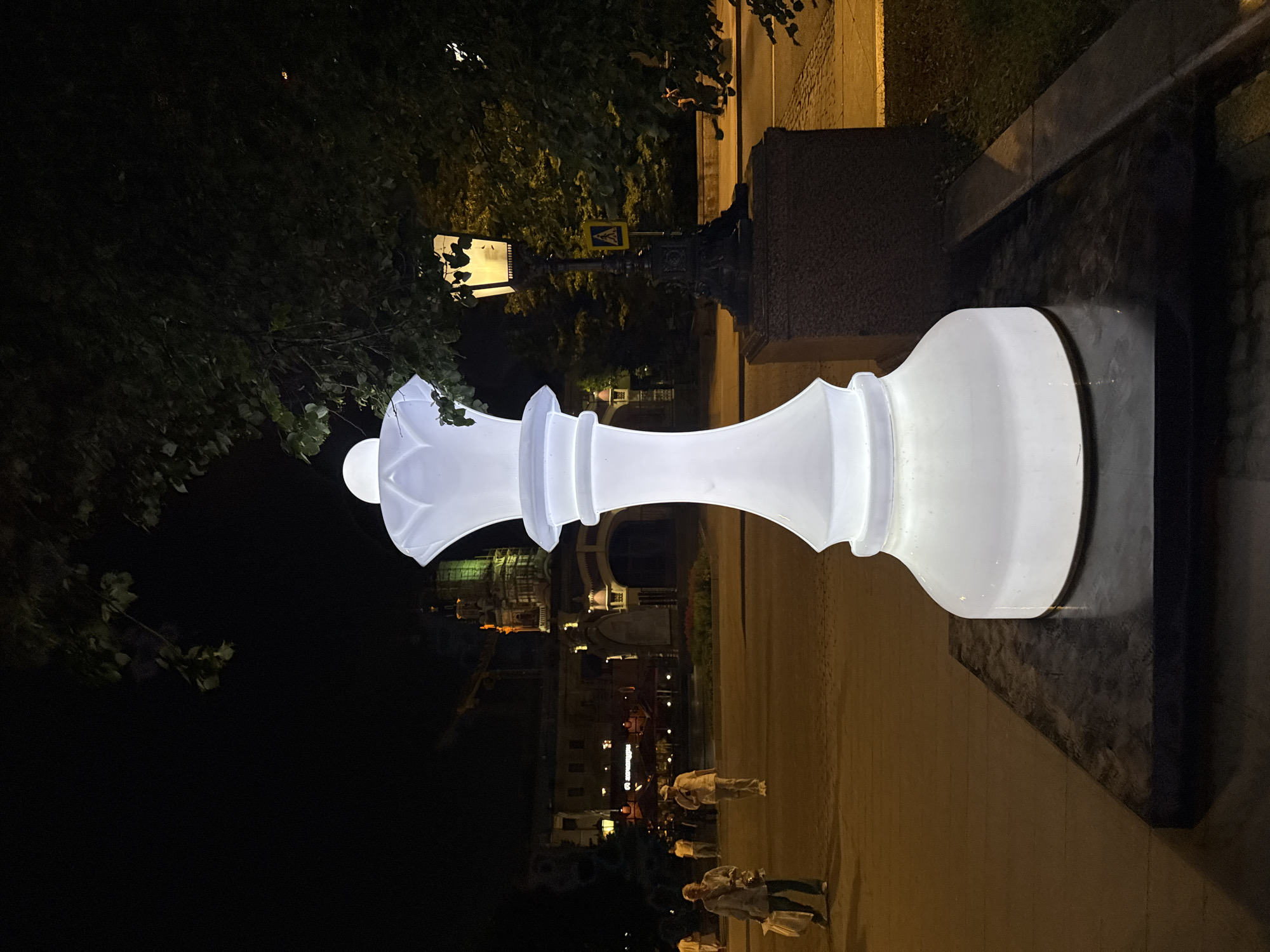 An illuminated, oversized white queen chess piece stands prominently in the foreground on a dark, rectangular base. The chess piece has a smooth, translucent surface that emits a soft glow. Behind it, a street lamp with an ornate dark metal base and a visible light source illuminates the surrounding area. A yellow pedestrian crossing sign is visible on the post of the street lamp.
In the background, several individuals are walking on a paved area. One person in the mid-ground is wearing light-colored clothing and carrying a bag. Another person to the left is also in light clothing and carrying shopping bags. Further back, a building with multiple windows and illuminated sections is visible, along with a crane and other structures suggesting an urban environment. The scene is at night, indicated by the darkness of the sky and the artificial lighting.
The setting appears to be a public space, possibly a plaza or park, within a city. Based on the architectural style of the background building and the presence of a crane, the location is identified as Moscow, Russia, likely near a landmark or a bustling area.