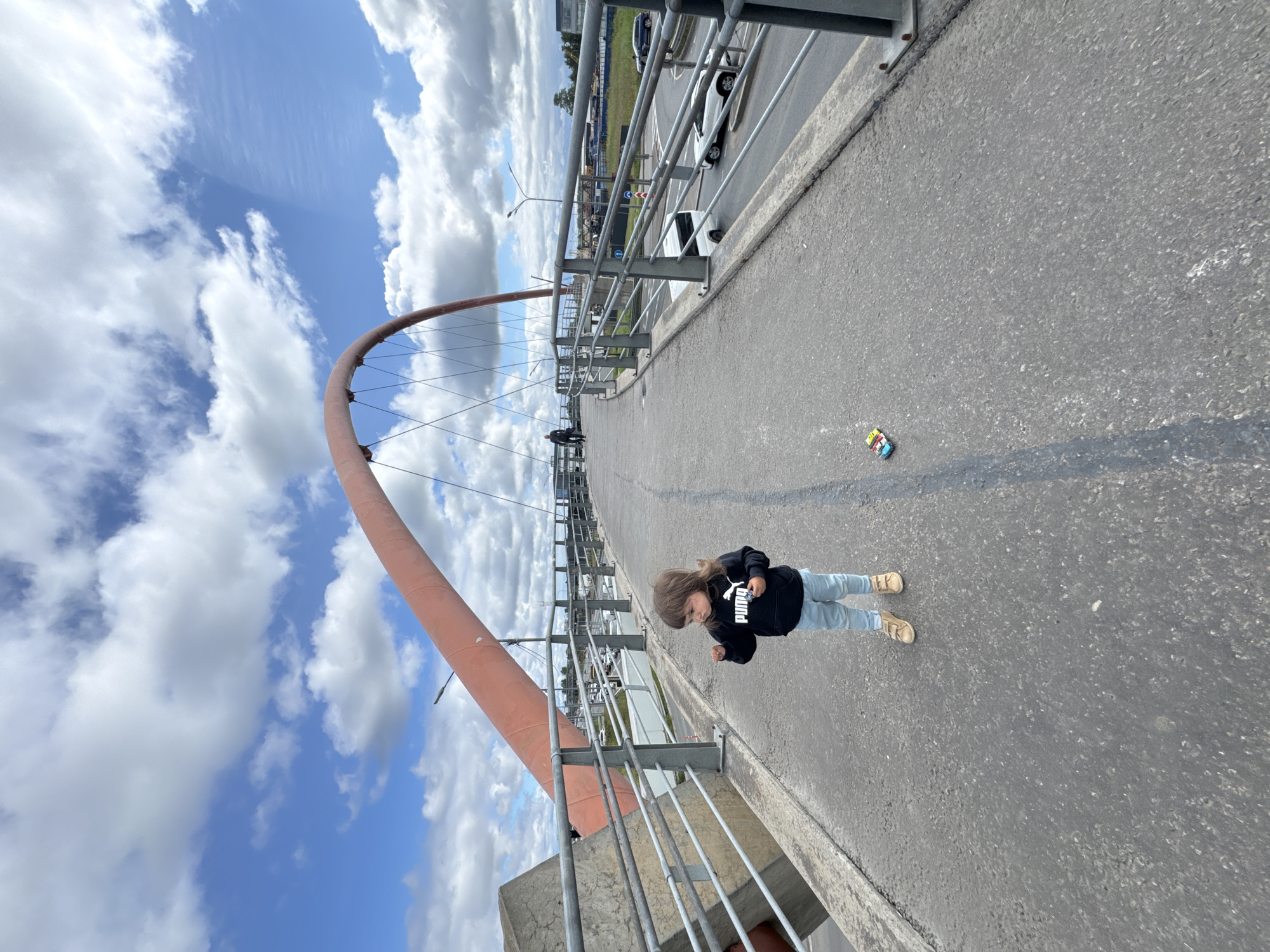 A young child stands on a pedestrian bridge in Saint Petersburg, Russia. The child is wearing a black Puma hoodie and light blue pants. A small toy car is on the ground near the child. The bridge has a red arch support and metal railings. In the background, cars are visible on a road below the bridge, and buildings and trees line the horizon. The sky is filled with white clouds against a blue backdrop. Another person is walking further down the bridge.