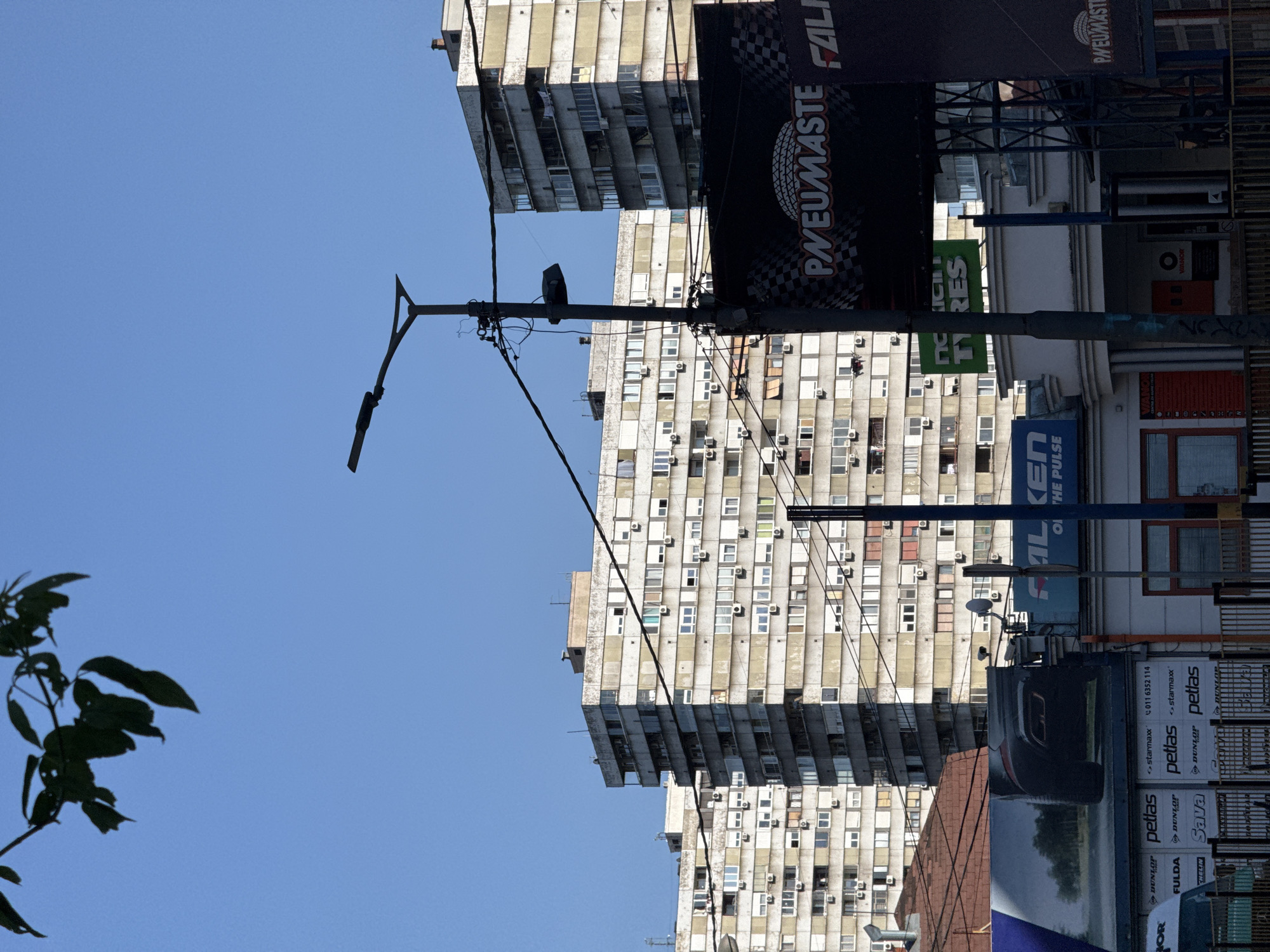 This media file captures an urban street scene dominated by tall, multi-story apartment buildings under a clear blue sky. The buildings feature numerous windows, almost all equipped with external air conditioning units, suggesting a residential or mixed-use purpose. Their concrete facades appear aged with some visible wear and tear.
A prominent street light pole stands centrally, with an array of electrical wires stretching across the frame. Several large commercial billboards are attached to structures adjacent to the buildings. A silhouetted branch with leaves occupies the lower left corner of the foreground.
The visible text on the billboards primarily advertises tire brands. Large black billboards prominently display "PNEUMASTE" with a checkered flag motif and "FALKEN," along with the tagline "ON THE PULSE." A green sign shows "TYRES," and another sign reads "VIANOR." Several smaller, stacked signs advertise "DUNLOP," "petlas," "starmaxx," "Sava," and "FULDA," accompanied by a phone number "011 6352 114."
The scene is set during the daytime under bright, sunny weather. There are no people visible, and no specific activity or event is taking place, presenting a static snapshot of an urban environment, consistent with a street in Vozdovac Urban Municipality, Serbia.