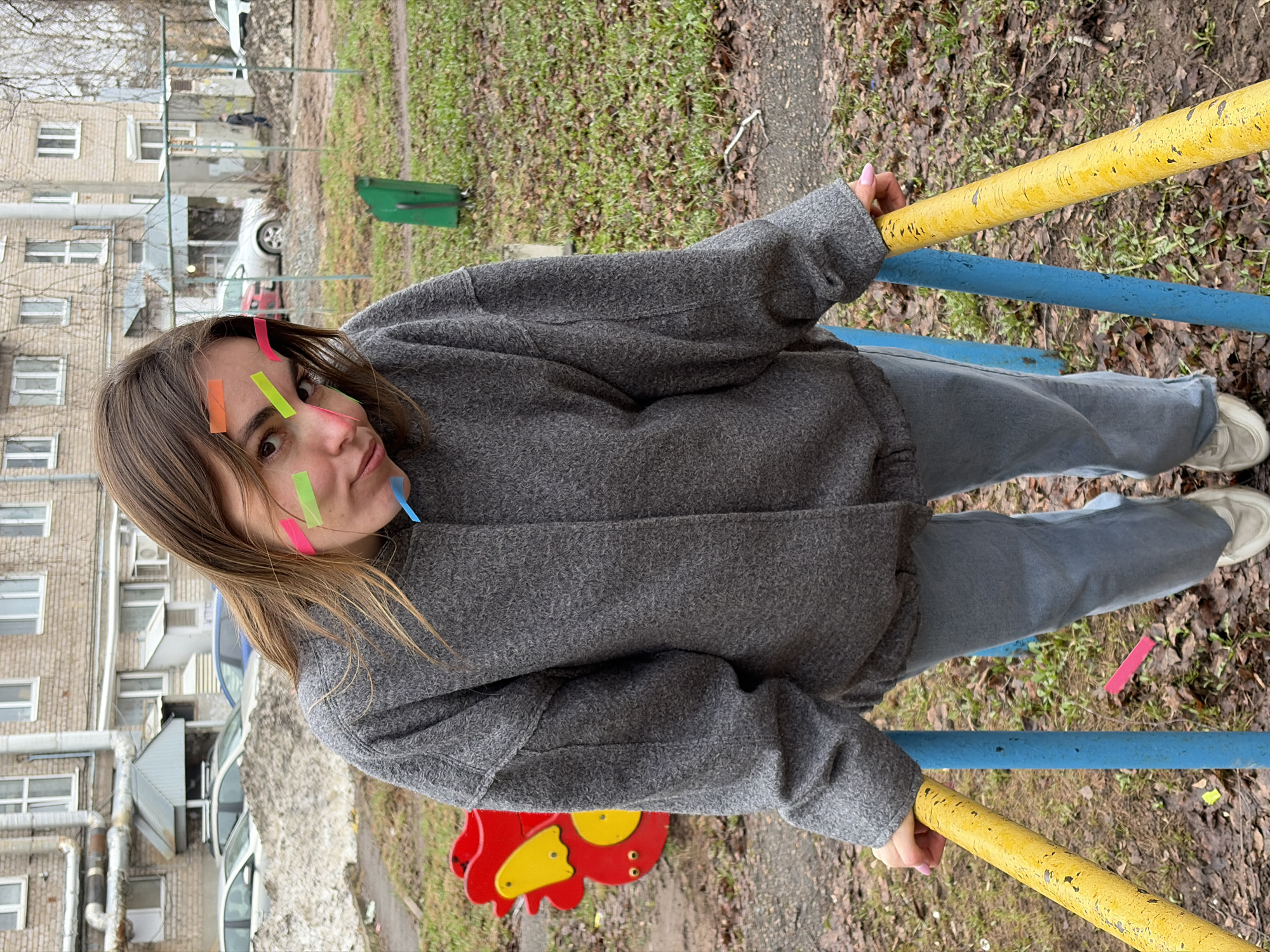 A woman stands near a playground in Izhevsk, Russia. She wears a gray jacket and pants and has colorful tape on her face. She holds onto yellow and blue bars, likely part of the playground equipment. The ground has grass, dirt, and some fallen leaves. An apartment building can be seen in the background. A red and yellow playground toy is visible as well. Cars are parked nearby.