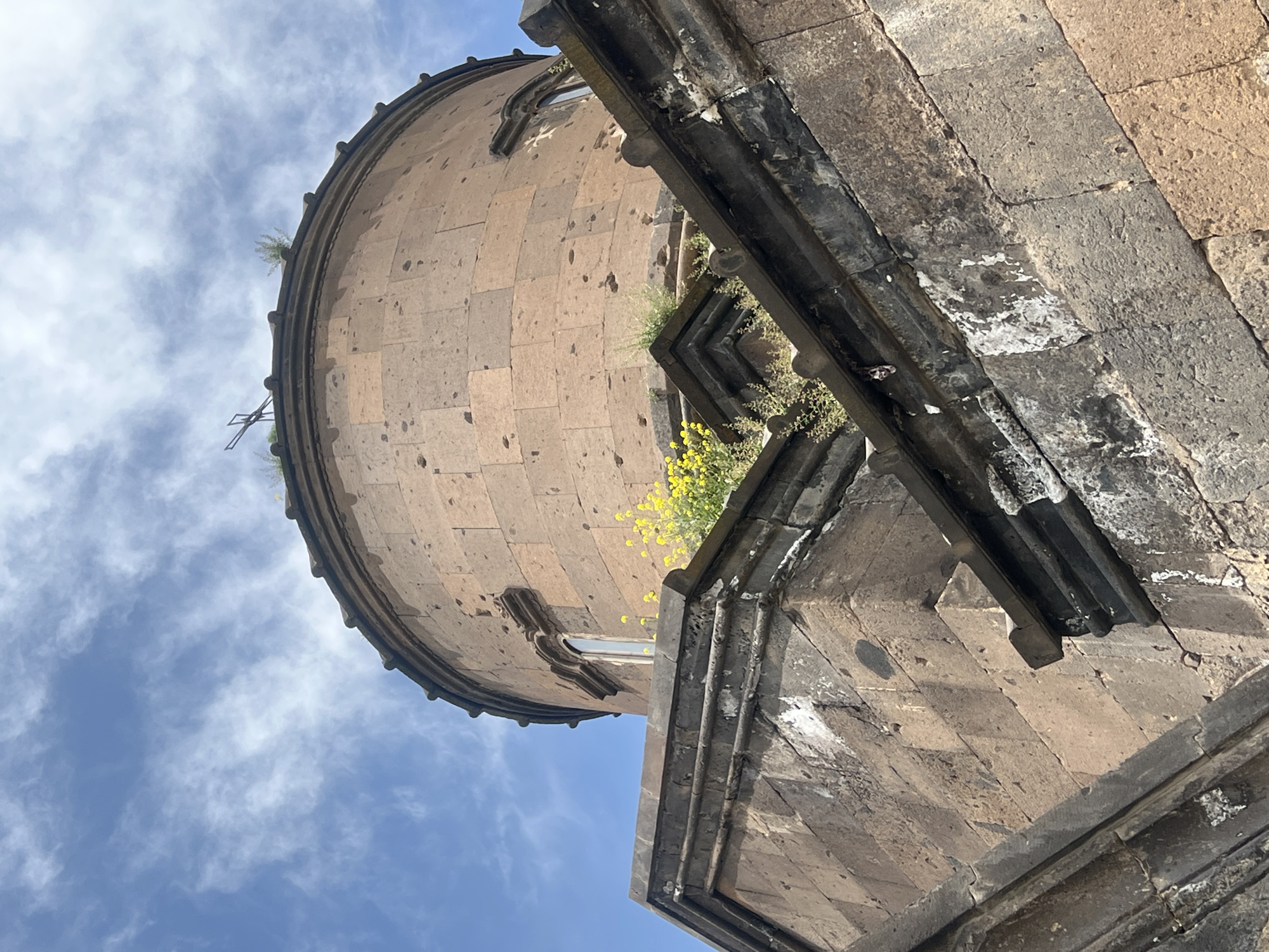 The image shows the dome of the Saint Sion Church, located in Artashavan, Armenia. The dome is constructed of light brown stone blocks and is round in shape. There are small plants growing on the dome and yellow flowers are visible on the adjacent stone structure. The sky is blue with scattered white clouds. A portion of the stone structure with dark grey accents is visible in the foreground.