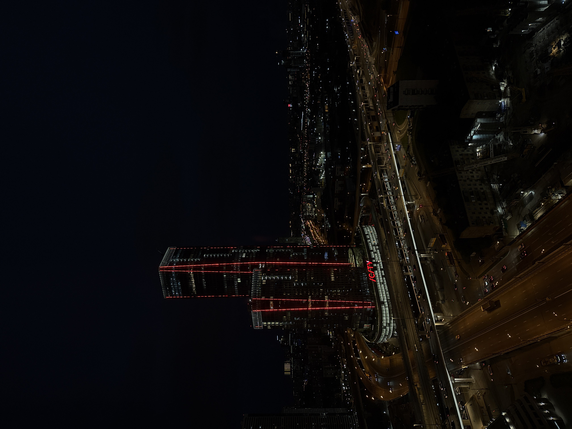 This is an aerial, nighttime photograph of the Moscow City business district in Khimki, Russia, oriented vertically. The scene depicts a sprawling urban landscape dominated by illuminated skyscrapers and a complex network of elevated highways.
The most prominent structures are two modern, glass-facade skyscrapers with striking red horizontal lighting that wraps around their exteriors. One of these buildings clearly displays the text "iCITY" in red neon letters. The surrounding area is densely populated with other buildings, many of which have illuminated windows, contributing to the city's glow.
A multi-lane elevated highway curves through the frame, carrying a visible stream of traffic. The headlights and taillights of the vehicles create streaks of light, indicating movement. In the lower right portion of the image, construction activity is evident, with cranes and partially built structures visible.
The time of day is clearly night, given the darkness of the sky and the reliance on artificial illumination. The weather appears clear, allowing for unobstructed views of the city lights and structures. The overall mood is one of urban dynamism and vastness, highlighting the scale and activity of a major metropolitan area at night.