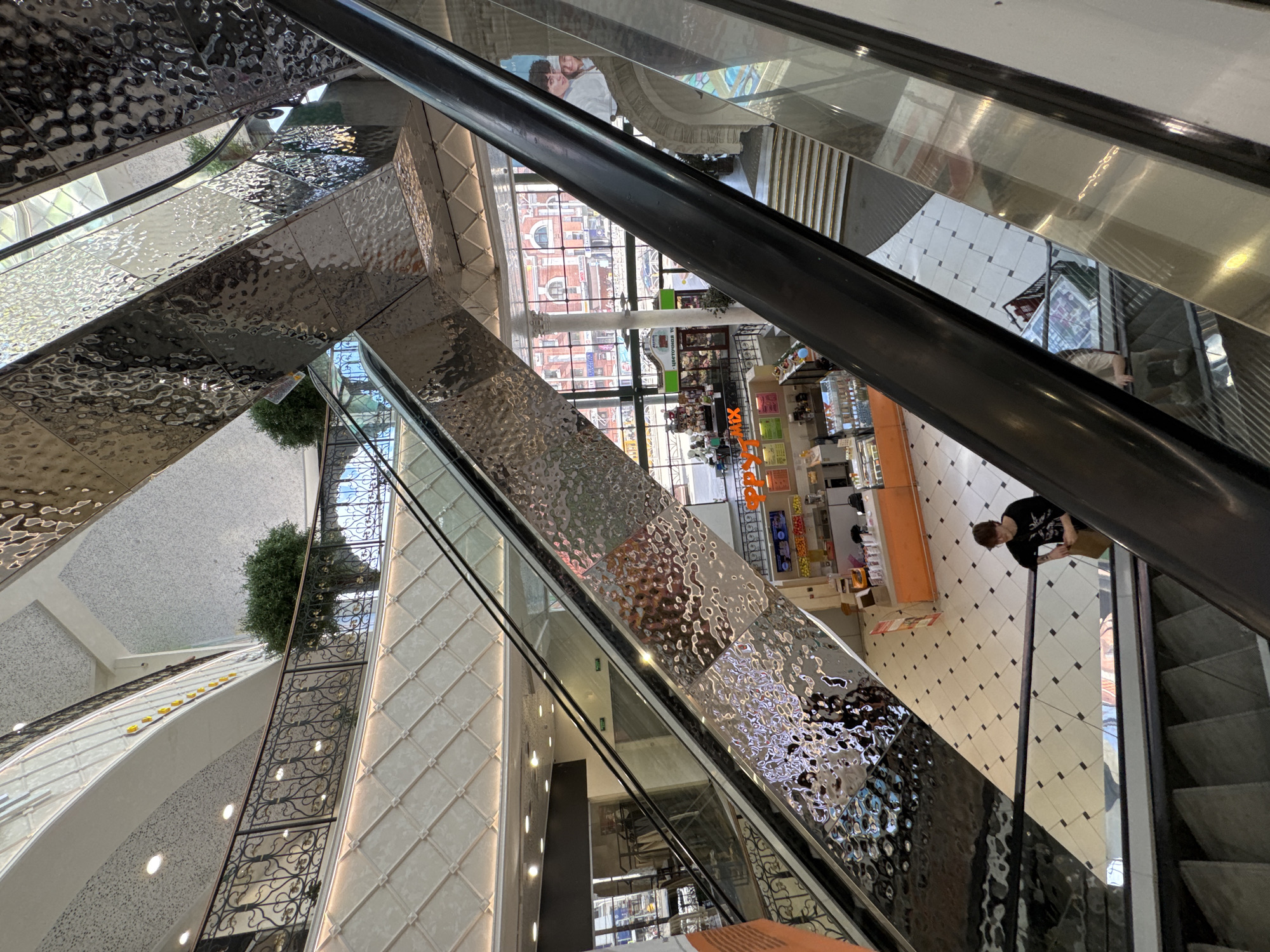 The image captures the interior of the "Greenwich" shopping mall in Yekaterinburg, Russia. The perspective is from an escalator looking downwards. The mall features multiple levels with escalators and walkways, decorated with decorative railings and textured metallic panels.
On the lower level, visible through the escalators and walkways, is a food court area with various stalls. One stall is brightly colored orange and yellow, with a sign that appears to say "HAPPY MIX". There are also tables and seating areas within the food court.
A young person wearing a black t-shirt and glasses is seen standing on the tiled floor of the mall, holding a brown paper bag. Another person is visible on an escalator further down, partially obscured.
The mall's architecture is modern, with a mix of white tiled floors, textured walls, and metallic accents. Large windows in the background reveal views of city buildings, including some with red brick facades. Artificial plants are strategically placed on balconies and ledges. Lighting fixtures are embedded in the ceilings. The overall impression is one of a bustling, multi-level retail and dining space.