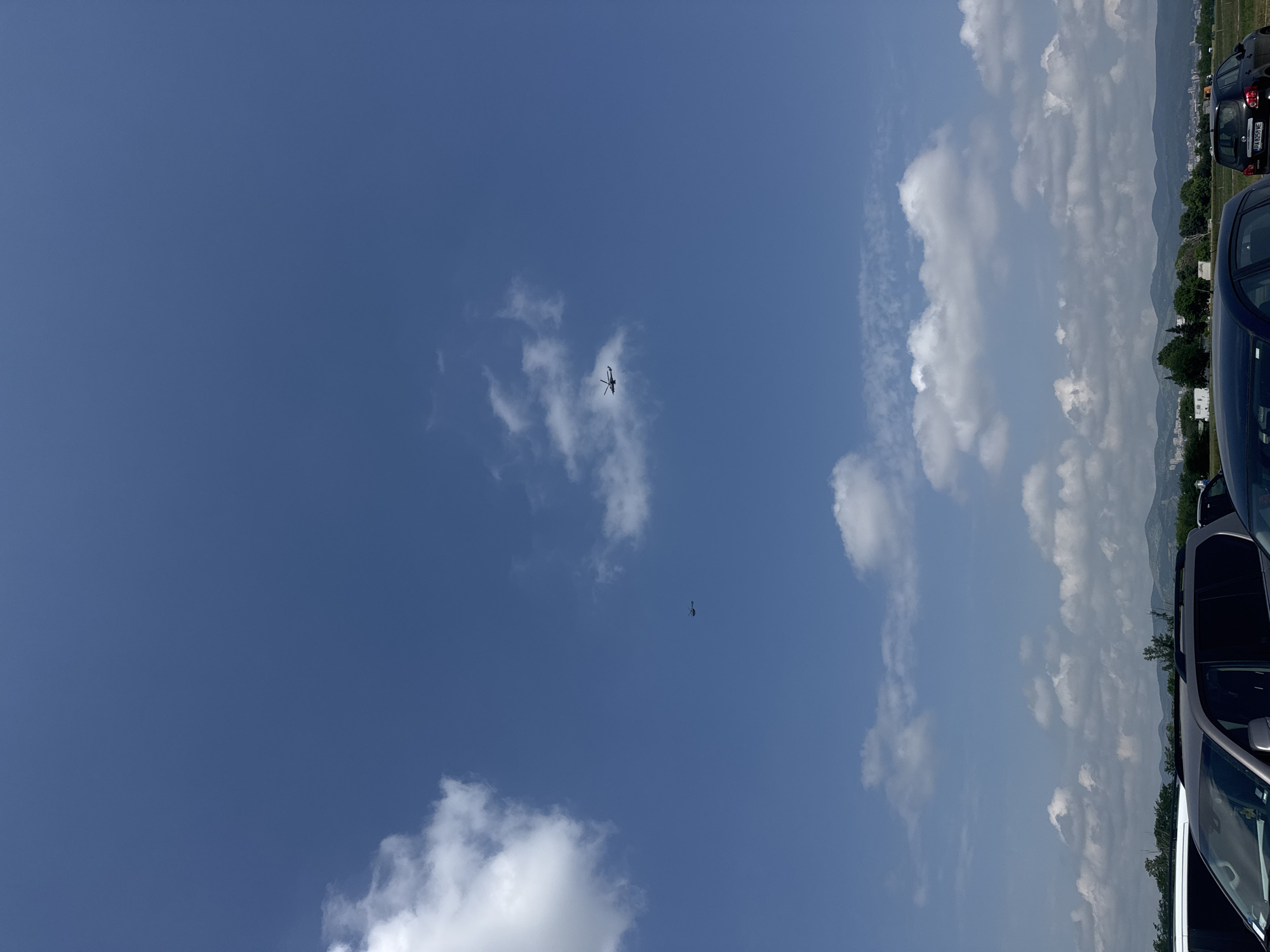 This vertically oriented photograph captures an open sky with aerial activity above a distant landscape, taken in Stara Zagora, Bulgaria. The upper portion of the image is dominated by a clear blue sky with scattered white cumulus clouds.
High in the sky, slightly to the left of the center, a dark silhouette of a helicopter is visible, showing its main rotor and tail boom against the bright sky. Below and to the right of the helicopter, a smaller, dark, elongated figure appears to be a person descending, consistent with a parachutist or skydiver, though the parachute itself is not distinctly open or visible.
The lower and right-hand side of the image features elements of a ground scene. Parts of several parked cars are visible along the right edge and in the bottom right corner. One dark car in the upper right has a partially visible license plate, which reads "CT 8546 HE," with "CT" being a regional identifier for Stara Zagora. In the background, a range of distant hills or mountains forms the horizon line, with some trees and what seem to be distant buildings at their base.
The time of day appears to be bright daylight, likely midday or early afternoon, given the clear sky and well-lit clouds. The weather is sunny with some cloud cover. The scene suggests an outdoor event featuring aerial displays or recreation, such as skydiving, observed from a parking lot or open area near a mountainous region.