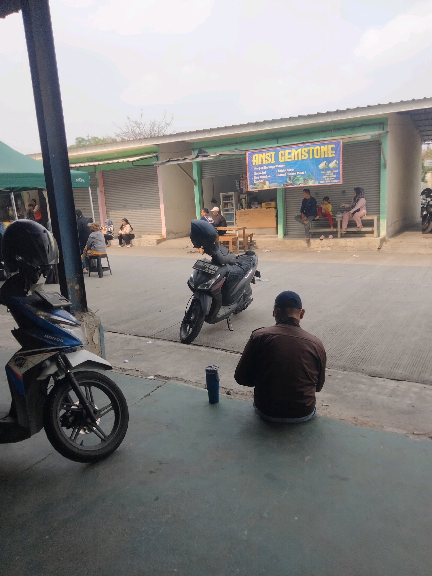 The image depicts an outdoor scene in front of a shop in Sukaasih, Indonesia. The shop has a large blue banner with the text "ANSI GEMSTONE" and images of what appear to be gemstones. Several people are visible in front of and inside the shop. In the foreground, two motorcycles are parked. A man wearing a brown jacket and a blue cap is sitting on the ground, facing away from the camera. Further back, near the shop, several individuals are seated on a bench or on stools, and some are standing. The environment suggests a street or marketplace setting with a building and paved ground. The sky is overcast.