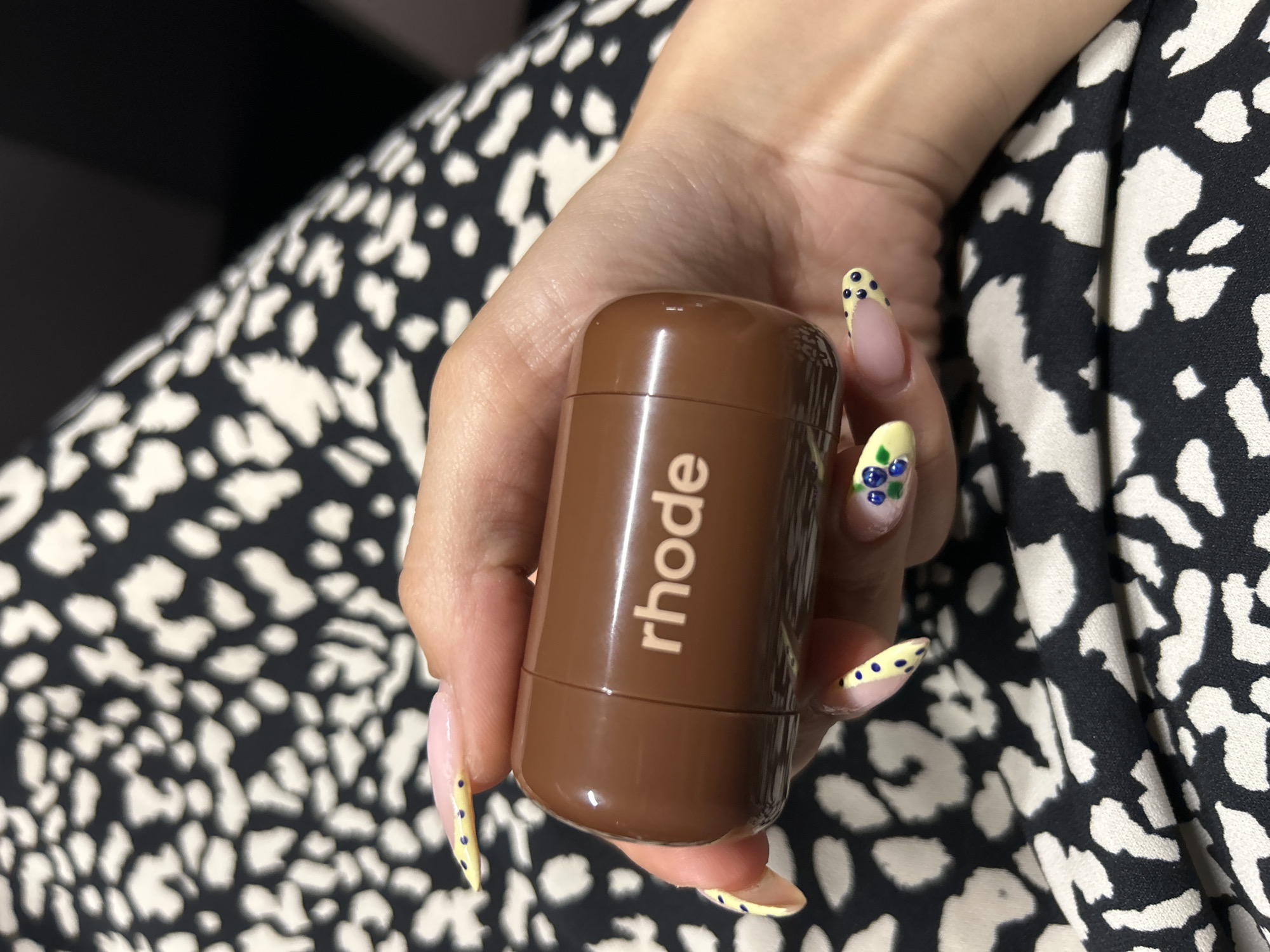 A person's hand, adorned with light pink and yellow nail polish decorated with blue dots and small blue flowers, holds a small, rounded, brown cylindrical object. The object has a light brown logo with the text "rhode" printed vertically on its side. The background is a black and white leopard print fabric, possibly a dress or a scarf, with some dark areas suggesting a dimly lit environment. The lighting appears to be soft and diffused, and there are no clear indicators of the time of day or weather. The scene is a close-up shot focusing on the hand and the object, with the background out of focus. The overall impression is one of personal care or beauty product display. No specific location cues beyond Tbilisi, Georgia are visible.
