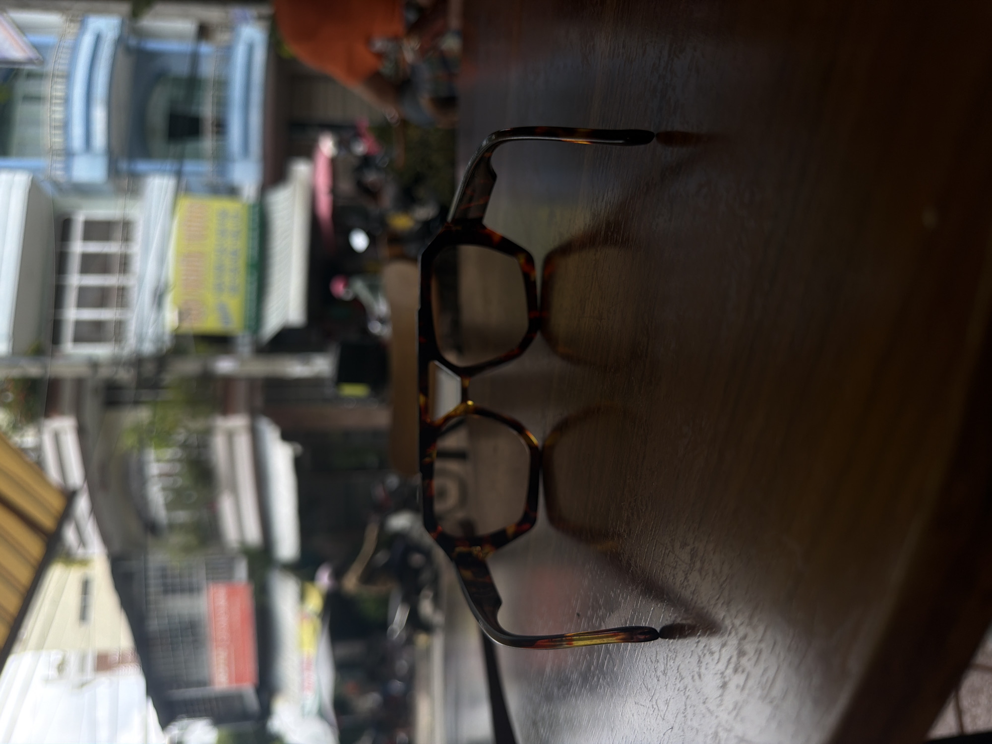 The image shows a pair of tortoise shell glasses resting on a wooden table in what appears to be an outdoor cafe setting. The background, slightly blurred, reveals a street scene in Nha Trang Ward, Vietnam, characterized by multi-story buildings, street signs, trees, and scooters parked along the road.