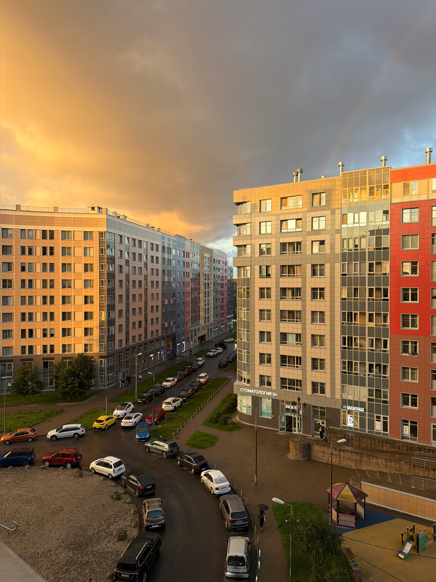 The image captures a street scene in Saint Petersburg, Russia, during a sunset with a dramatic sky and a faint rainbow. Multiple modern, multi-story apartment buildings with colorful facades dominate the frame. The buildings are densely packed, with a variety of window styles and architectural details. A street runs alongside the buildings, with numerous parked cars of various makes and colors. The road appears wet, suggesting recent rain. There are streetlights and some greenery, including trees and a small playground area with a colorful structure. A few people are visible on the sidewalk, one holding an umbrella. The overall impression is of an urban environment bathed in the warm, golden light of the setting sun, contrasted by dark, cloudy skies.