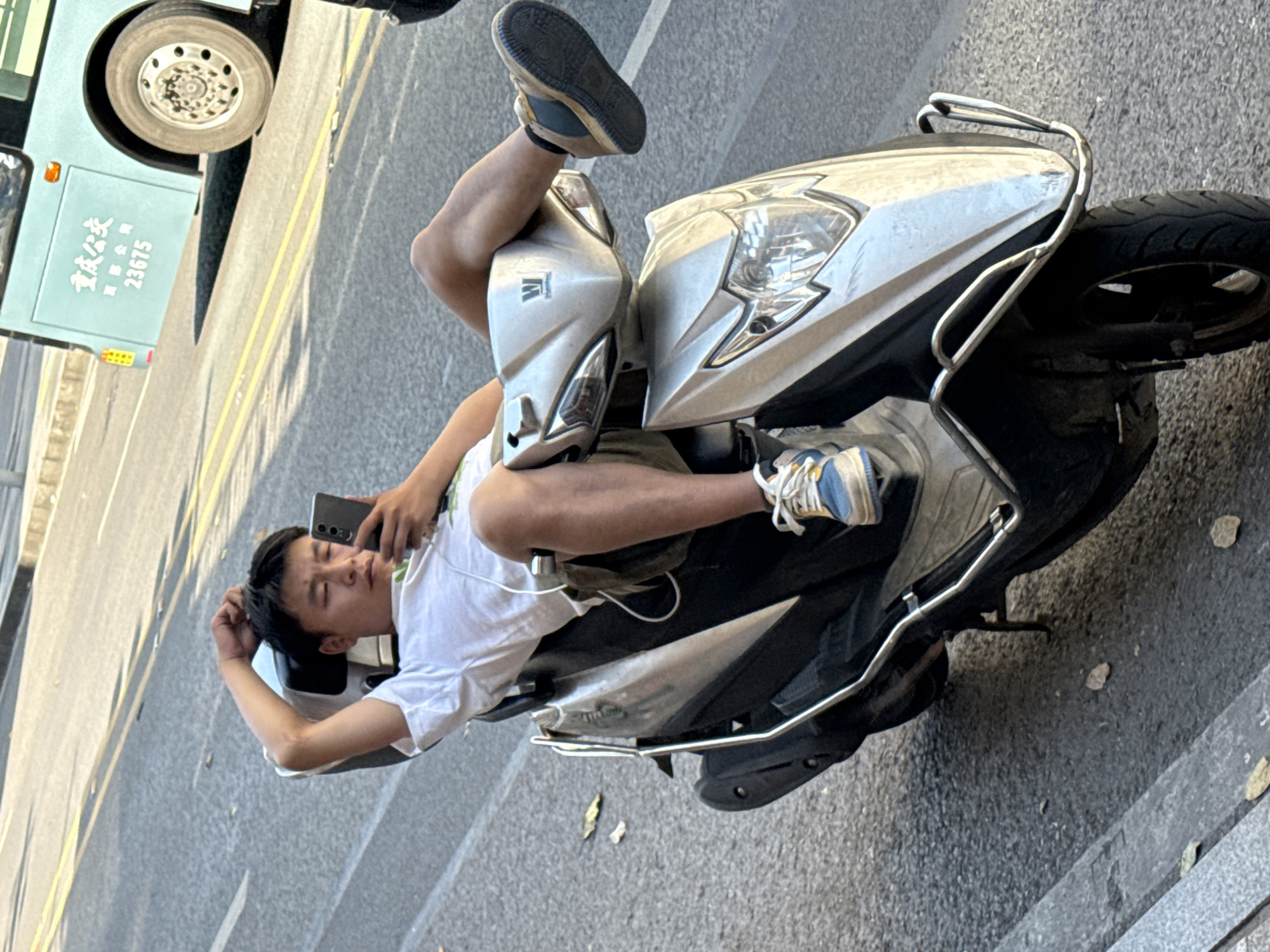 A young Asian man is lying prone on a silver scooter on a city street. He is wearing a white t-shirt and shorts, and one foot rests on the scooter's front. The other leg is elevated in the air. The man is holding a smartphone in one hand. A bus is visible parked next to the sidewalk. The text on the bus reads '重慶公交集团' (Chongqing Public Transportation Group) '23675'. The location is likely in the Jiulongpo District in Chongqing, China.