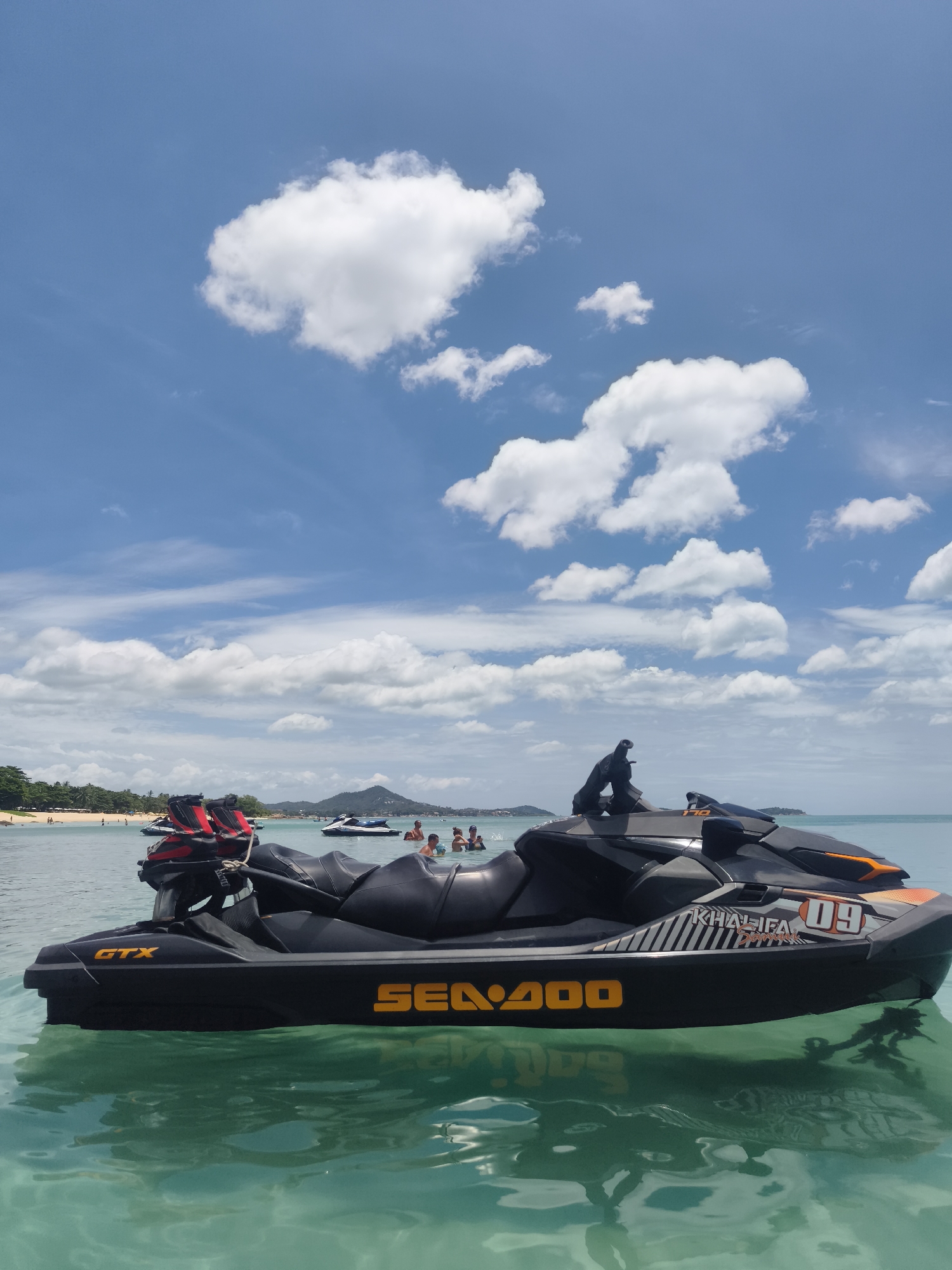 In the clear turquoise waters off the coast of Ko Samui District, Thailand, a black Sea-Doo GTX jet ski floats serenely. The jet ski is positioned in the foreground, with its sleek design and orange accents visible. The water is so transparent that the underside of the jet ski is partially visible, and reflections of the sky and the vessel shimmer on the surface.
In the background, the sandy beach of Ko Samui is lined with lush green palm trees, hinting at the tropical paradise. Several other jet skis and a few people are scattered in the shallow water, enjoying the sunny day. In the distance, a few boats are anchored, and a mountainous silhouette of the island rises against a backdrop of a bright blue sky dotted with fluffy white clouds. The overall scene evokes a sense of leisure and fun in a beautiful coastal environment.