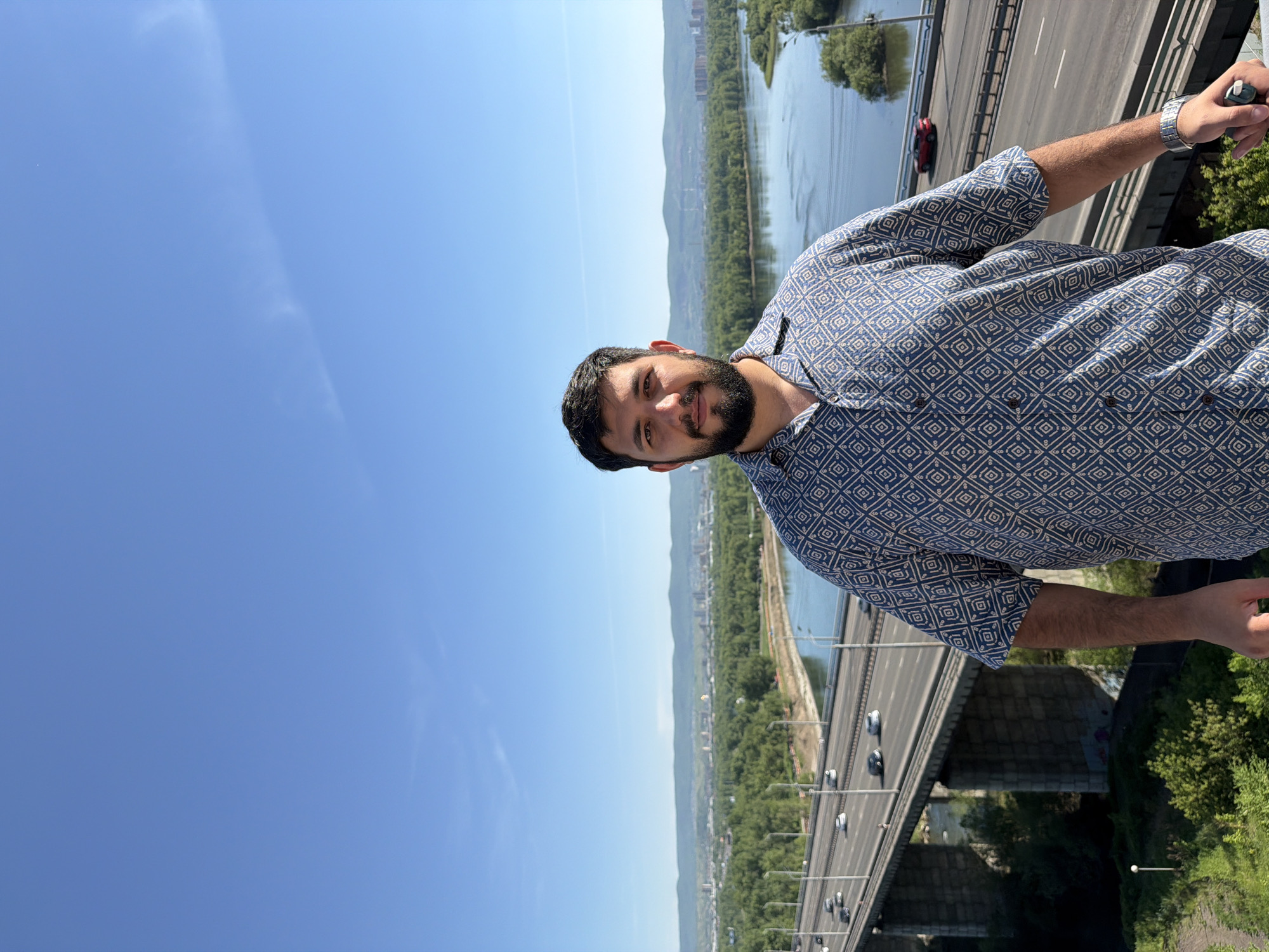 The image depicts a male individual positioned in the foreground, facing the camera directly, against an expansive outdoor backdrop of Krasnoyarsk, Russia. The man has dark hair, a full beard, and a mustache. He is wearing a short-sleeved collared shirt with a blue base and a repeating geometric pattern of light-colored diamond shapes. A silver-toned wristwatch is visible on his left wrist, and his right hand grasps a dark object, possibly a camera or phone, resting on an unseen railing.
The background reveals a wide, calm river, consistent with the Yenisei River, flowing from the left to the right of the frame. A multi-lane concrete bridge, likely one of Krasnoyarsk's major crossings such as the Fourth Bridge or Oktyabrsky Bridge, spans the river, carrying vehicular traffic. Several cars are visible on the bridge, some blurred due to motion. Dense green foliage and trees line the riverbanks and surround the bridge's concrete support pillars. In the mid-distance, a city skyline with various buildings is discernible, backed by a range of rolling hills or low mountains. Industrial structures, potentially factory chimneys, are visible in the distant hilly landscape to the right. The sky is clear and bright blue, indicative of daylight and fair weather, with only a few faint wisps of clouds present in the upper right quadrant. The foreground shows green vegetation, suggesting the photograph was taken from an elevated vantage point overlooking the city and river.