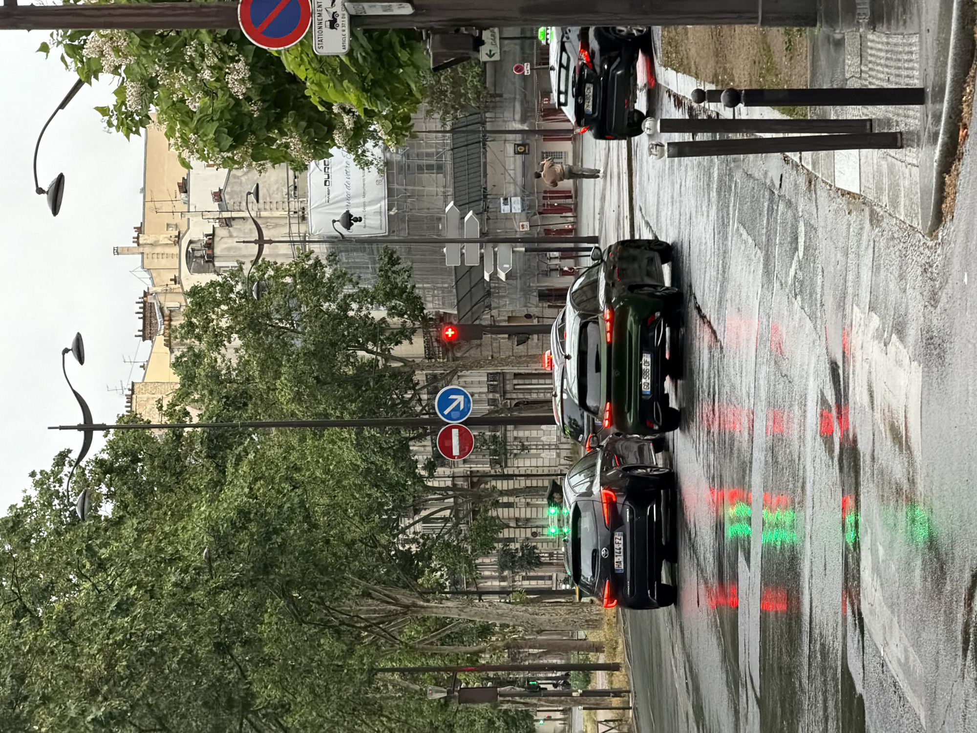The image captures a street scene in Paris, France, on an overcast day, likely after or during light rain, as evidenced by the wet asphalt and prominent reflections. The view looks down a tree-lined street with multi-story, classic Parisian buildings on either side.
In the foreground and middle ground, several cars are queued on the wet road, appearing to be stopped at a traffic light or in heavy traffic. Their brake lights (red) are illuminated, and vivid reflections of red and green traffic lights create streaks of color on the glistening road surface. Visible license plates include "HA-144-FZ", "GO-586-OF", and "FH-480-CN", consistent with French registration.
Along the street, various road signs are posted on poles. These include a "No Parking" sign (red circle with blue slash), a "No Entry" sign (red circle with white bar), and a "Compulsory Direction - Straight Ahead" sign (blue circle with white arrow). Another sign reads "STATIONNEMENT" (Parking) and "ARTICLE R 37.1 DU CO", indicating local parking regulations. In the background, an advertisement or construction sign partially reads "DURE" and "service de votre c", with "GARA" visible nearby.
The buildings lining the street exhibit traditional Parisian architecture, featuring light-colored stone facades, multiple windows, and chimneys. One building in the distance is partially covered by scaffolding, suggesting ongoing construction or renovation. Lush green trees with white blossoms line the street, partially obscuring the buildings. A single person, dressed in a light jacket, is visible walking or standing further down the street near the construction area. Overhead streetlights with decorative fixtures are also present.