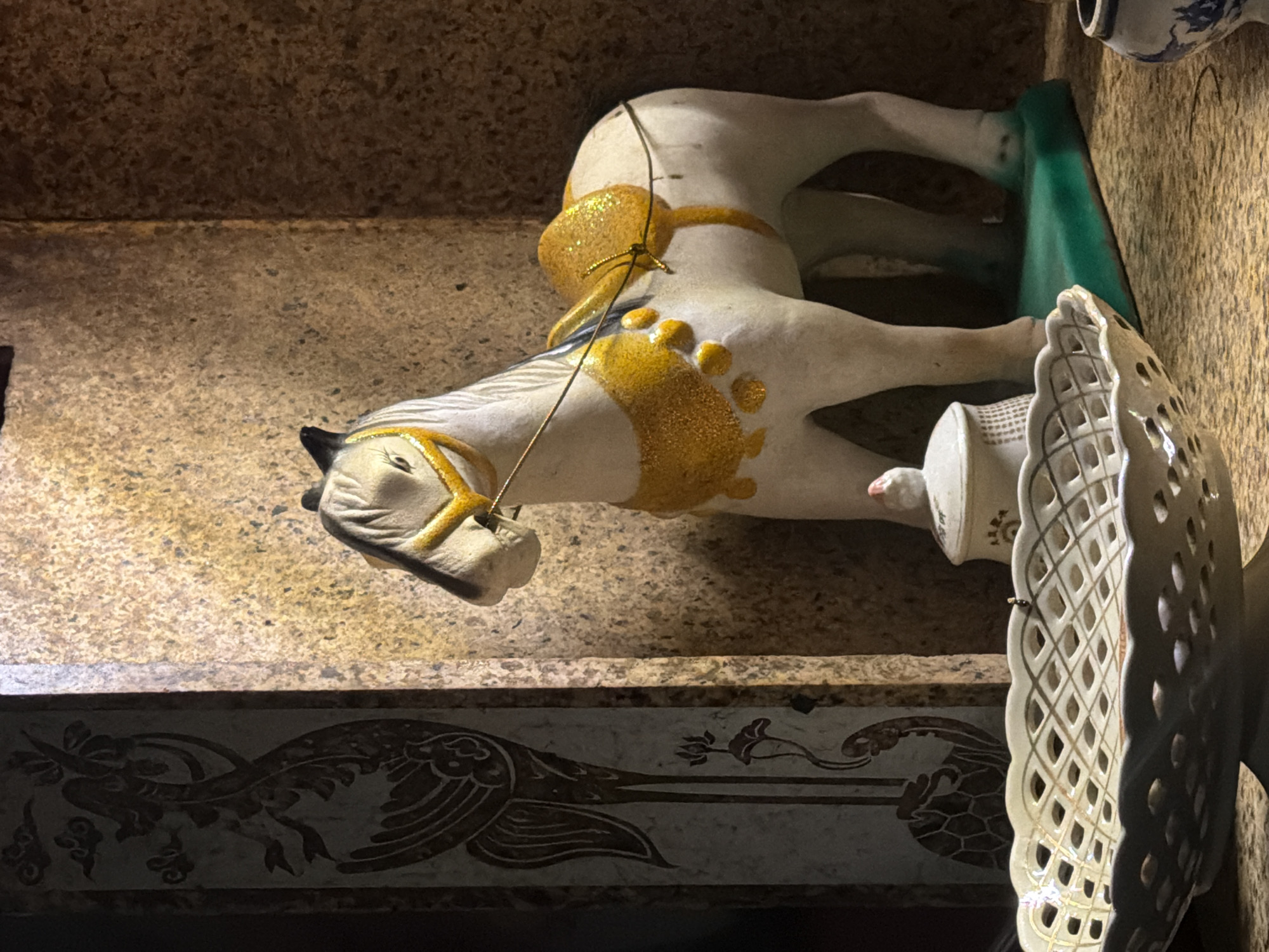 This media file presents a static composition of decorative objects, likely displayed on a shelf or ledge within an indoor setting. The central and most prominent subject is a white figurine of a horse, oriented on its left side. This ornamental horse is extensively decorated with shimmering gold glitter, outlining its mane, forming a saddle-like pattern on its back, and accentuating its legs or hooves. A thin wire is subtly attached to its back, suggesting it might have been hung or part of a larger arrangement.
To the right of the horse, a decorative white ceramic or porcelain plate lies on its side, showcasing an intricate openwork or lattice pattern, possibly featuring delicate gold accents. Partially visible behind the horse’s back is a small, bright green object. In the upper right corner, a fragment of a blue and white patterned ceramic item, potentially a vase or jar, is also discernible.
Below the main display, a distinct decorative panel or border adorns a wall or lower surface. This panel features elaborate dark brown or bronze-colored motifs set against a lighter, possibly off-white, background. The designs include a stylized bird, resembling a phoenix or peacock, with prominent, detailed wings, intertwined with various floral and vine-like patterns. The surrounding surfaces, both above and below the objects, are textured and mottled in shades of brown and beige, suggesting stone or concrete. The overall lighting is somewhat dim, creating subtle shadows. The arrangement evokes a sense of cultural display or artistry, commonly found in a home, shop, or temple in Đà Nẵng, Vietnam. No human figures are present, no specific activity is taking place, and no text is visible in the image.