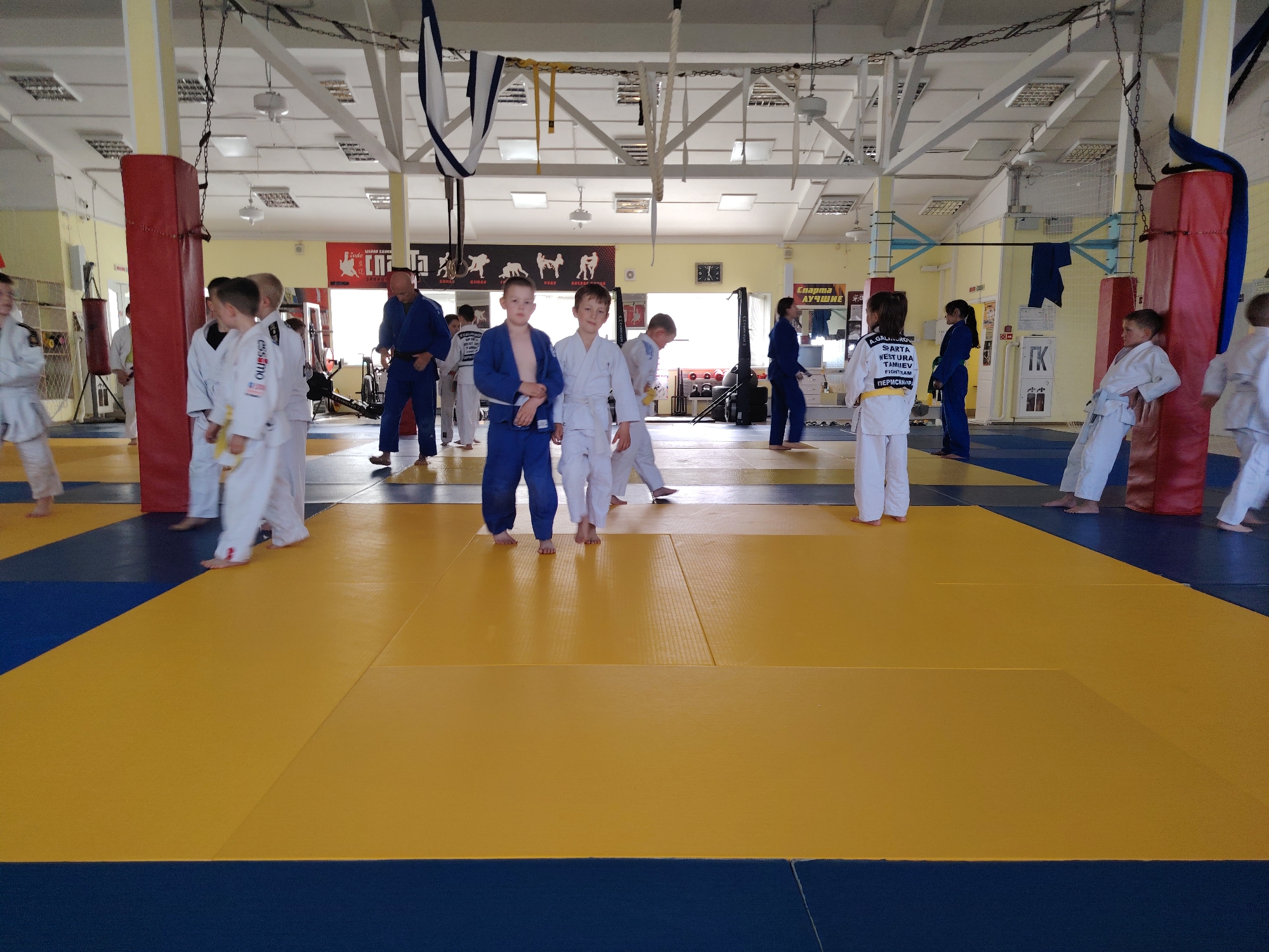 The image shows a group of children and adults in a judo hall in Chaikovsky, Russia. The hall has a large judo mat with yellow and blue sections. There are support columns, some wrapped in red protective padding. In the background are posters in Russian script on the wall and various exercise machines. Several individuals are wearing judo uniforms, some blue and some white. The group seems to be preparing for or practicing judo.