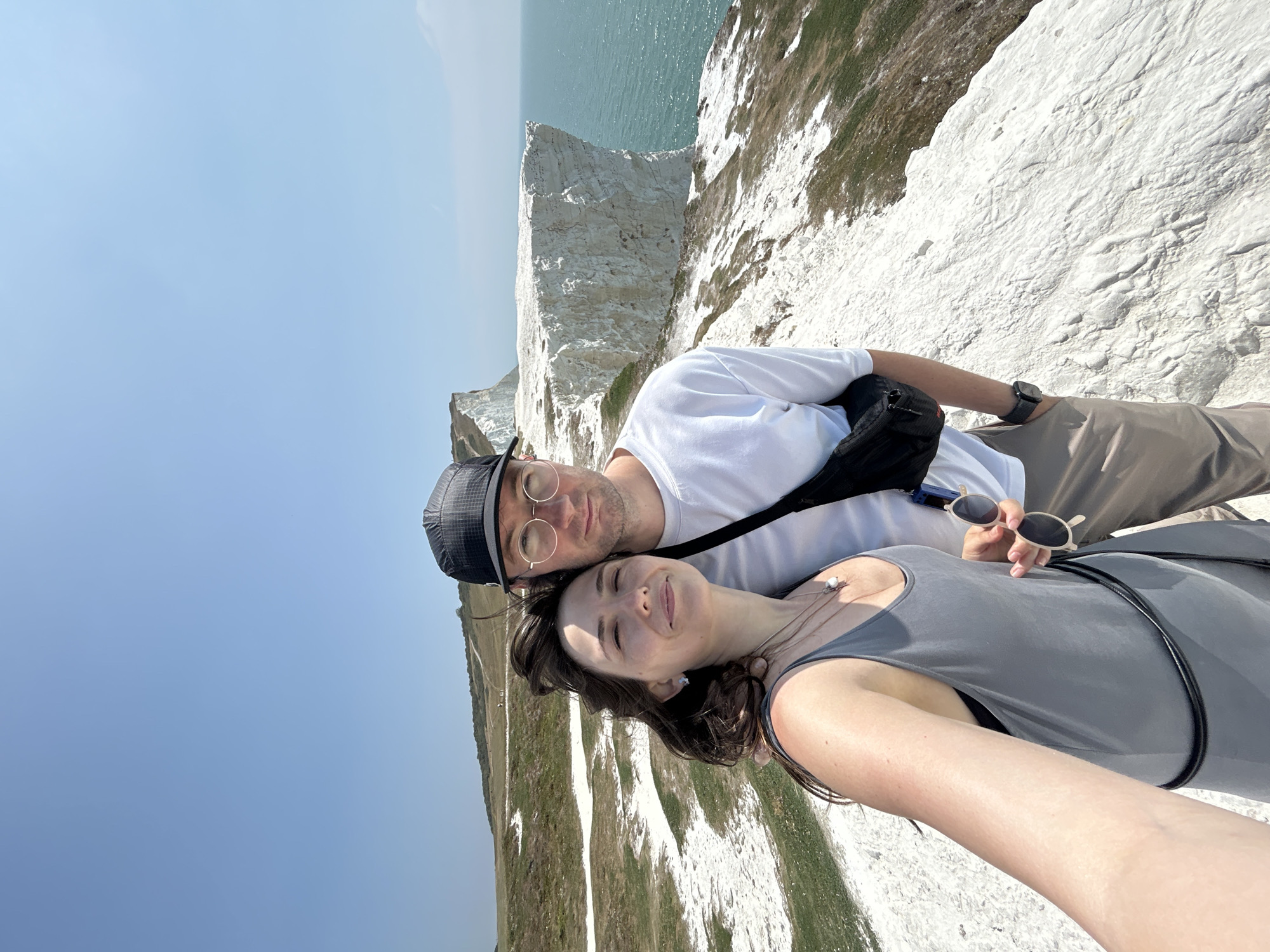 A couple is captured in a selfie on a cliff edge overlooking the ocean in Seaford, United Kingdom. The woman, on the left, is wearing a gray tank top and necklaces. The man, on the right, is wearing a white t-shirt, a black fanny pack, and a black baseball cap. They are both smiling at the camera. The background shows a vast expanse of white cliffs and the turquoise ocean. The sky is clear and blue. The ground is covered in chalky white material and sparse green grass. In the distance, other chalk cliffs and a pathway are visible. The sea is calm.