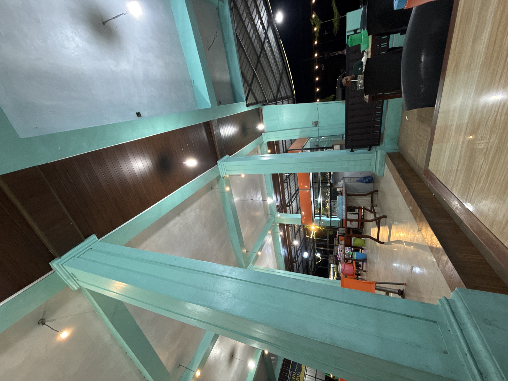 This media file captures an interior view of what appears to be a restaurant or cafe in Grong-Grong, Indonesia, at night. The scene is dominated by large, light blue painted wooden beams and columns, creating an open, airy architectural structure. The ceiling is a light color, possibly concrete or plaster, with visible lighting fixtures casting a warm glow.
The main subjects are the architectural elements and the ambient lighting. There is one person visible in the background, a man sitting at a table, appearing to drink from a straw. He is wearing headphones and a dark shirt. The setting includes tables and chairs, with some chairs being brightly colored (orange, green, blue) in the mid-ground. The floor is tiled with a wood-grain pattern. In the far background, through a glass partition, more tables and lighting are visible, suggesting a larger dining area.
Notable details include the artificial lighting, indicating it is nighttime. The overall mood is relaxed and inviting, typical of an evening dining establishment. The specific location cues are limited to the mentioned architectural style and the Indonesian context provided.
No discernible text is clearly visible in the image, though a sign that might contain text is partially visible in the far background.