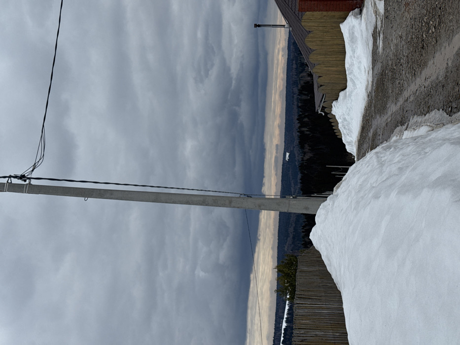 The image depicts an outdoor, rural or suburban scene in what is identified as the city of Sibir', Russia, under heavily overcast conditions. In the immediate foreground, a significant mound of white snow occupies the lower left, bordering a dirt or gravel path that appears partially thawed. Another, smaller snowdrift is visible on the right side of this path, adjacent to a wooden picket fence with a distinctive scalloped top edge.
A tall, grey concrete utility pole stands prominently in the center-left of the frame, extending from the bottom almost to the top, with several dark electrical wires attached and stretching horizontally across the upper midground. To the right of the path and wooden fence, a portion of a building with reddish-brown brick or siding and a dark, possibly tiled, roof featuring a metal chimney is visible.
The background presents an expansive landscape of densely forested hills, appearing dark and dense under a somber sky. A narrow, lighter band is discernible at the distant horizon line, separating the dark forest from the dominant, unbroken expanse of dark grey, cloudy sky. On one of the far-off forested hills, a distinct white, rectangular, horizontal object or feature is observed. No individuals or active subjects are present in the scene.