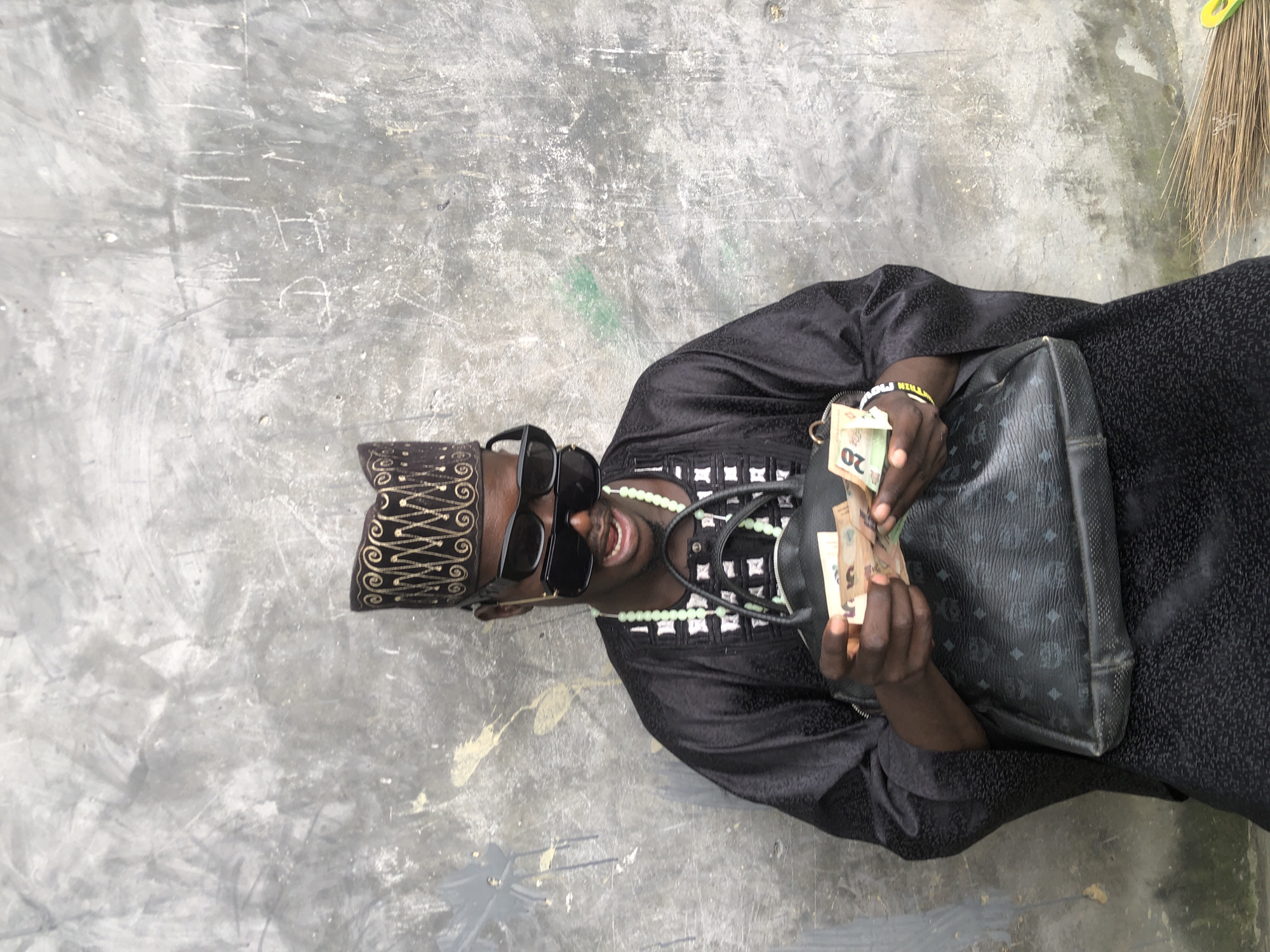 A person wearing a black, embroidered traditional outfit and a patterned cap is seated against a textured concrete wall. They are also wearing black sunglasses and a light green beaded necklace. The person is holding several banknotes in their hands, and a large black handbag with a repeating logo pattern is resting on their lap. The concrete wall appears aged, with some scuff marks and graffiti. A broom is visible in the lower right corner of the image. The lighting suggests it is daytime.