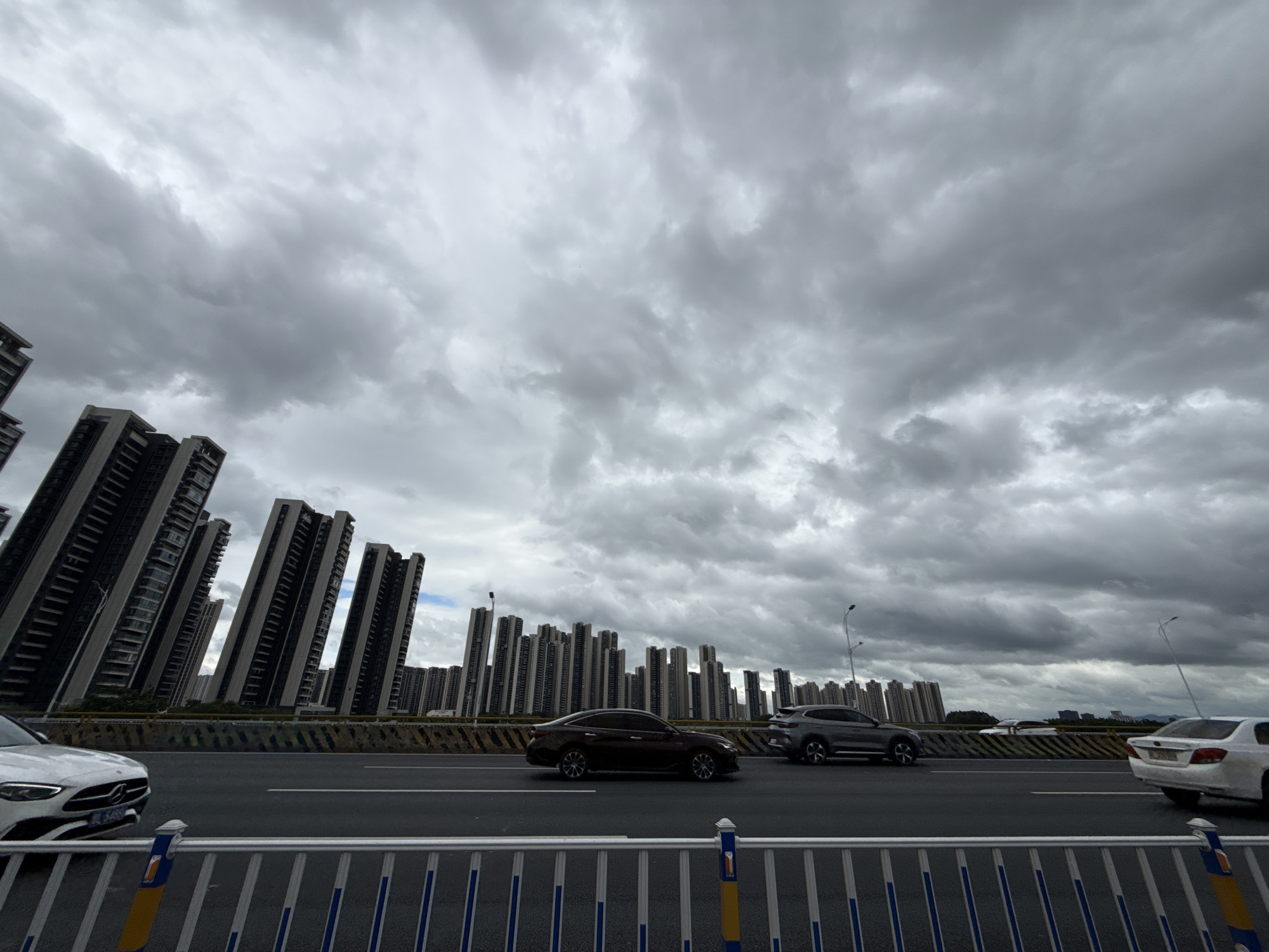 This media file captures a wide-angle view of a city skyline under a dramatic, cloudy sky. The scene features a multi-lane highway in the foreground, with several cars in motion. A white Mercedes-Benz is visible on the left, partially cropped. To its right, a dark-colored sedan is driving, followed by a grey SUV. Further down the road, a white sedan is seen from the rear on the right side of the frame.
Dominating the background is a dense collection of tall residential buildings, appearing to be apartment complexes. Many of these buildings are angled, giving the impression of leaning or being constructed on a slope, which is a notable detail in the composition. The sky is filled with dark, heavy clouds, suggesting overcast or potentially stormy weather. The overall lighting is diffused, indicative of either early morning, late afternoon, or a heavily clouded day.
The setting appears to be an urban environment in Huizhou, China, as suggested by the architecture and the urban infrastructure like the highway and the tall residential blocks. There is no visible text in the image. The mood evoked is one of urban sprawl under a somber, cloudy sky, with the angled buildings adding a unique, almost surreal visual element.