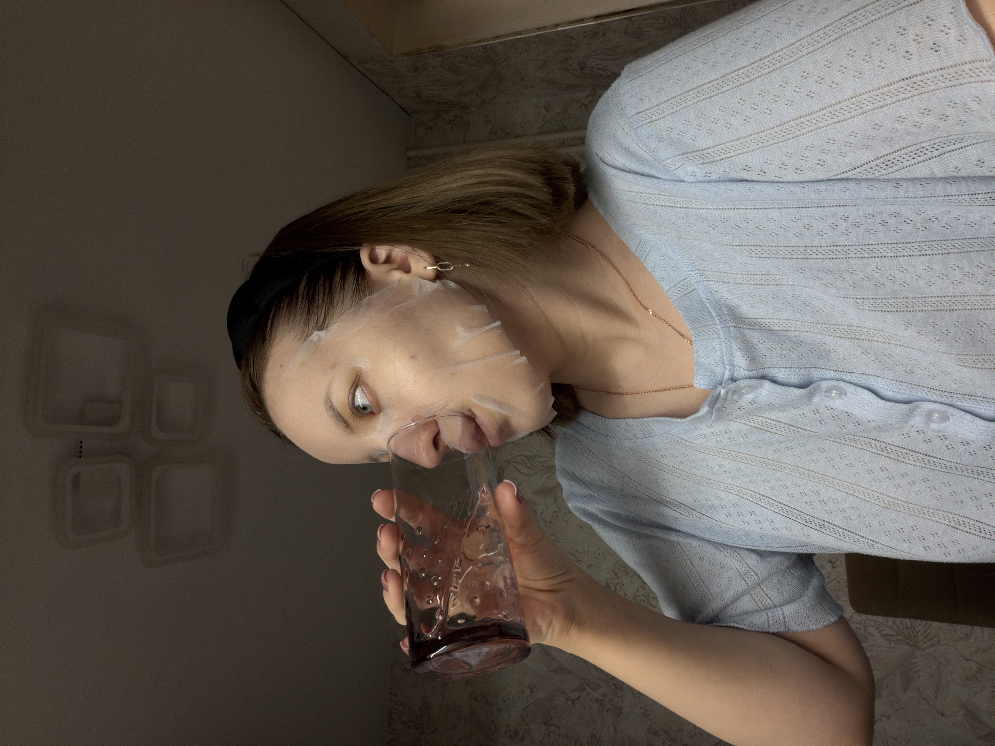 A woman is depicted drinking from a glass while wearing a face mask in an indoor setting. She has light brown hair, held back by a black headband, and her face is covered by a translucent white sheet mask, allowing her light-colored eyes to be visible as she looks towards the top left. She is wearing a light blue, semi-sheer, open-knit top or cardigan with a patterned texture and visible buttons, along with a thin gold necklace. Her right hand holds a textured, clear glass with a reddish-brown base, which she is using to drink a clear liquid. The background is dimly lit, featuring a patterned wall behind her and a darker wall area to her left with white, square-shaped decorative elements. The scene suggests a relaxed, self-care activity, likely in the evening. No text is visible in the image. This scene takes place in Reshetnikovo, Russia.
