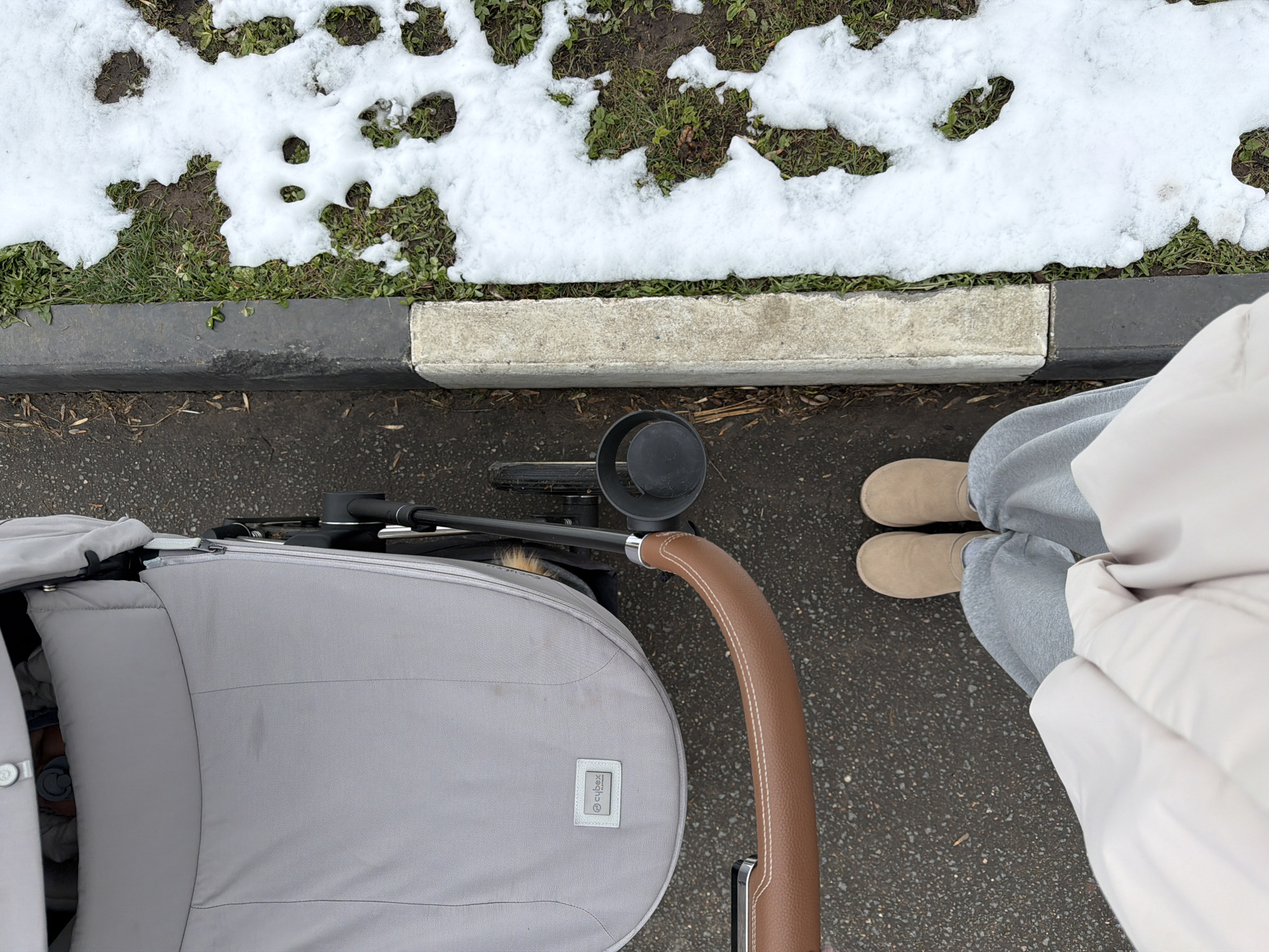 An overhead photograph captures a person standing beside a light grey baby stroller on an asphalt surface, with snow-covered grass and a curb in the background. The scene is set outdoors, likely on a sidewalk or path in Khimki, Russia.
The person's lower body is visible on the right, wearing light grey sweatpants and light brown, possibly suede, boots. They are also wearing a light-colored, presumably padded, jacket, part of which is seen. The large baby stroller, featuring a brown, stitched, leather-wrapped handle, occupies the left side of the frame. A small white rectangular tag with the "Cybex" brand name is visible on the side of the stroller's main body.
The ground directly under the person and stroller is dark, textured asphalt. To the left, a concrete curb separates the asphalt from a strip of ground covered in green grass and irregular patches of melting snow. The curb itself is painted in alternating sections of black and off-white. The presence of snow indicates cool or cold weather. The time of day is daylight. No specific activity beyond standing or walking with the stroller is evident, and no explicit emotions are discernible.