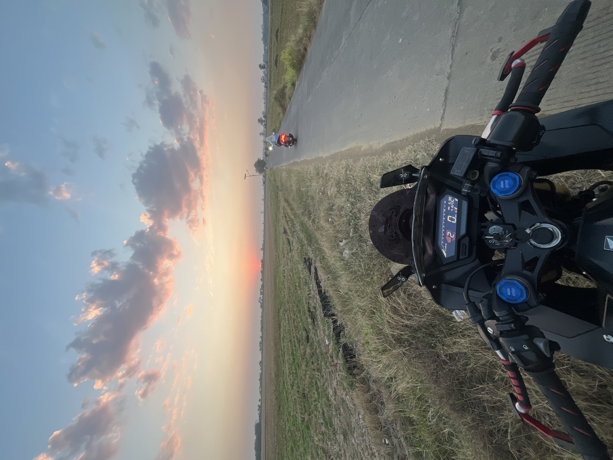 The image depicts a first-person perspective of someone riding a motorcycle on a paved road in a rural area, likely in or around Pecangaan, Indonesia, during sunset. The sky is a gradient of light blue and soft orange, with scattered clouds tinged pink by the setting sun. The road stretches ahead, bordered by dry, grassy fields. In the distance, another motorcycle is visible, also traveling on the road. The foreground shows the handlebars and instrument panel of the motorcycle being ridden, displaying a digital speedometer reading "0" and "5.38" with "41643" below it. The scene conveys a sense of movement and the peaceful atmosphere of a rural sunset.