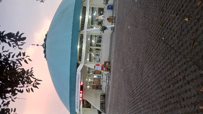 The image displays the exterior of Masjid Raudhaturrahmah in Pasar Paloh, Indonesia, during the evening. A large, light-blue dome with a finial crowns the white-walled mosque, which features multiple illuminated arched windows and openings. In the foreground, a dark gray interlocking paver surface shows sparse grass and scattered dry leaves. The main entrance, accessed via a short staircase, is marked by an overhead sign reading "MASJID RAUDHATURRAHMAH". An electronic display below the sign shows "19:30 WIB. A" and "Asr". To the left of the entrance, a red stand with a white sign and a small wooden stool are positioned near dark-colored benches. To the right, potted plants are visible alongside yellow and blue trash receptacles. Silhouetted tree foliage partially obscures the upper dome against a sky displaying gradients of pink, orange, and light blue. No individuals are discernible in the scene.