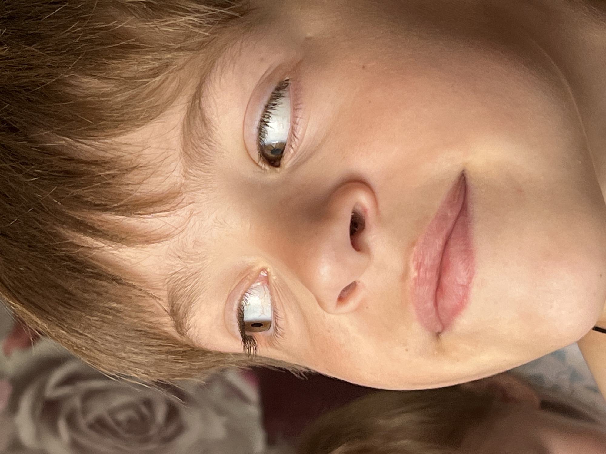 A close-up, eye-level portrait captures a young child with light brown hair and fair skin, located in Noviy, Russia. The child's brown eyes are directed sharply to the viewer's left and right, with visible eyelashes and slight redness in the sclera. The nose is small and upturned, and the lips are full and slightly parted. Scattered faint freckles are discernible across the cheeks and nose. A portion of another individual's head, out of focus, is visible on the far left. The background consists of a blurred, muted floral pattern, indicative of an indoor setting with patterned wallpaper. The lighting is soft and natural, illuminating the child's face from the front-right.