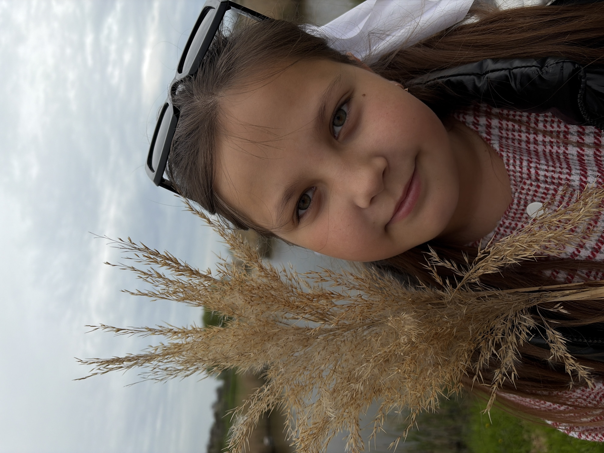 A young girl with brown hair and green eyes is holding a bouquet of dried reeds. She is wearing a red and white patterned dress and a black jacket. Sunglasses are perched on her head, and a white bow is in her hair. The background appears to be an outdoor scene, possibly near a body of water, under a cloudy sky. The location is Novopokrovskaya, Russia.
