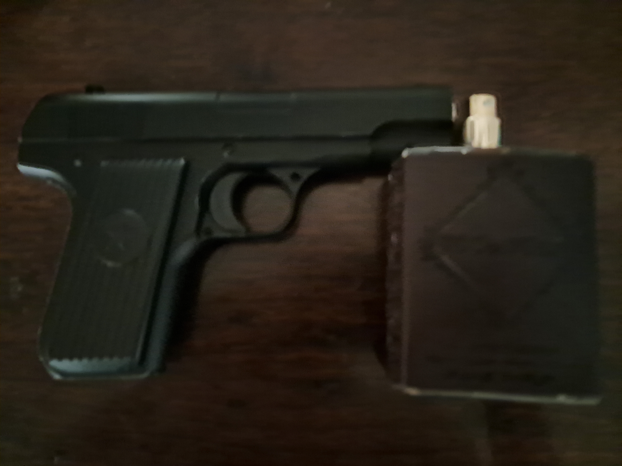 The image shows a black handgun resting on a dark wooden surface. Below the handgun, there is a dark rectangular object, possibly a magazine or a case. The handgun is positioned vertically, with its barrel pointing upwards. The rectangular object is placed horizontally beneath the grip of the handgun. The wooden surface appears to have a visible grain. The lighting in the image is dim, and the overall focus is soft, giving a somewhat blurry appearance. There are no people visible in the image, and no specific location in Bagish, Uzbekistan, can be identified.