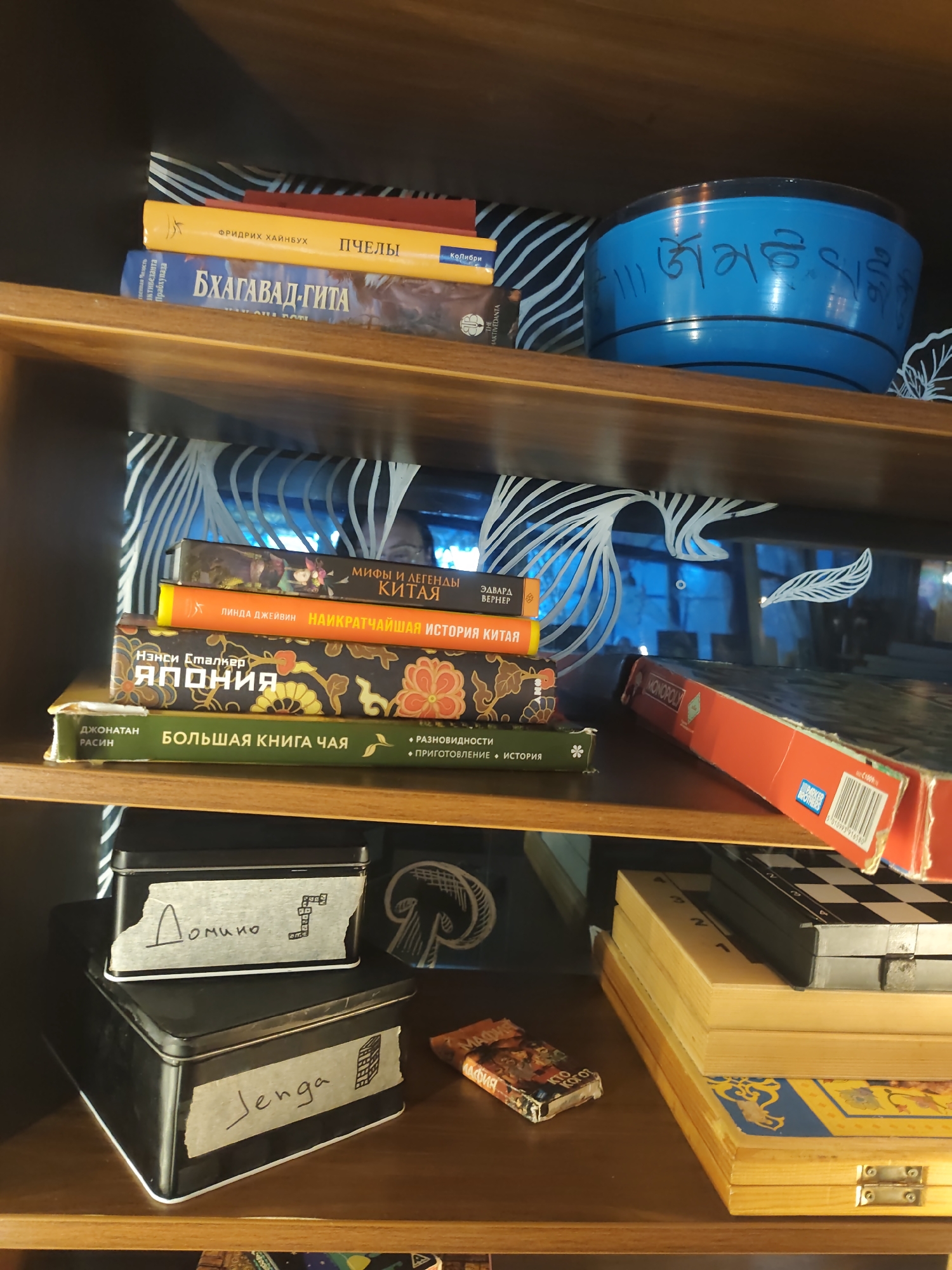 The image depicts a wooden shelf in what appears to be a room, possibly a home or a shop, in Sochi, Russia. On the top shelf, several books are stacked, with a blue bowl adorned with Tibetan script to their right. Below this, another shelf holds more books, including ones with titles that appear to be in Russian and possibly other languages. A red Monopoly box lies on this shelf. The bottom shelf contains two black metal boxes with labels, one marked "Домино" (Domino) and the other "Jenga." A small, colorful box with Russian writing is also present on this shelf. A chessboard and other board game boxes are visible on the far right. The overall setting suggests a collection of books and games.