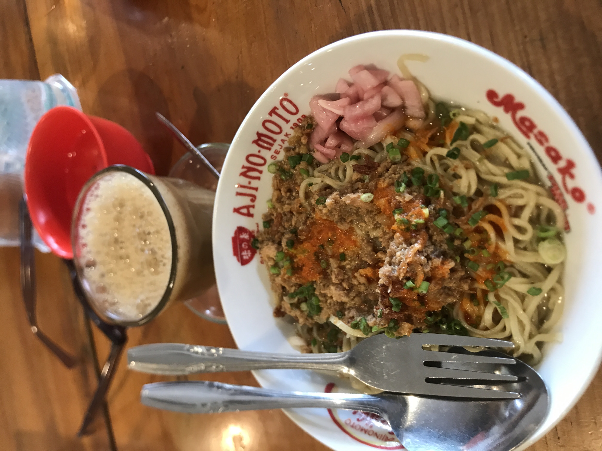 The image depicts a bowl of Mie Ayam Masako, a chicken noodle dish, in Bandarbireuen, Indonesia. The noodles are topped with seasoned chicken, pickled vegetables, and scallions. A fork and spoon rest on the bowl's rim. A glass of foamy beverage sits beside the dish. A red plastic cup and clear container are also visible. The scene is set on a wooden table.