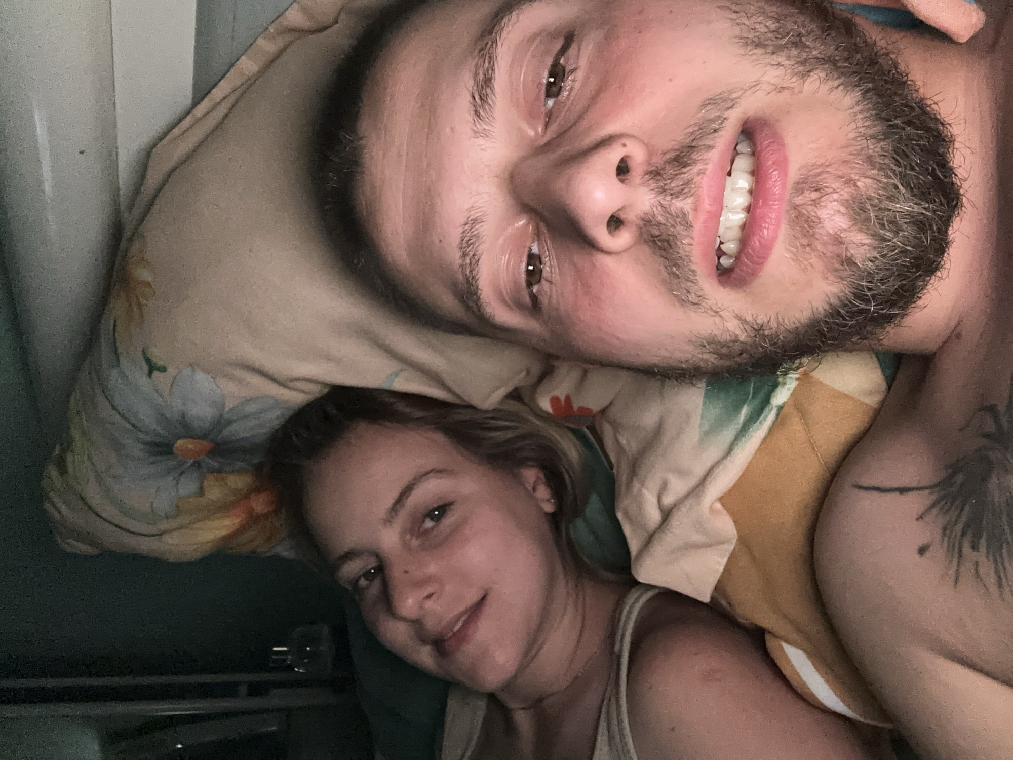 An overhead, indoor, close-up selfie shows a man and a woman lying in bed. The man is in the foreground, his face filling most of the frame from the nose down. He has dark hair, a beard, and is smiling with his teeth showing. The woman is behind him, to his left, her head resting on a pillow. She has blonde hair and is looking at the camera with a slight smile. Both individuals are wearing tank tops. The background consists of a pillow with a floral pattern and a dark green wall. The lighting is dim, suggesting it is nighttime or a dimly lit room.