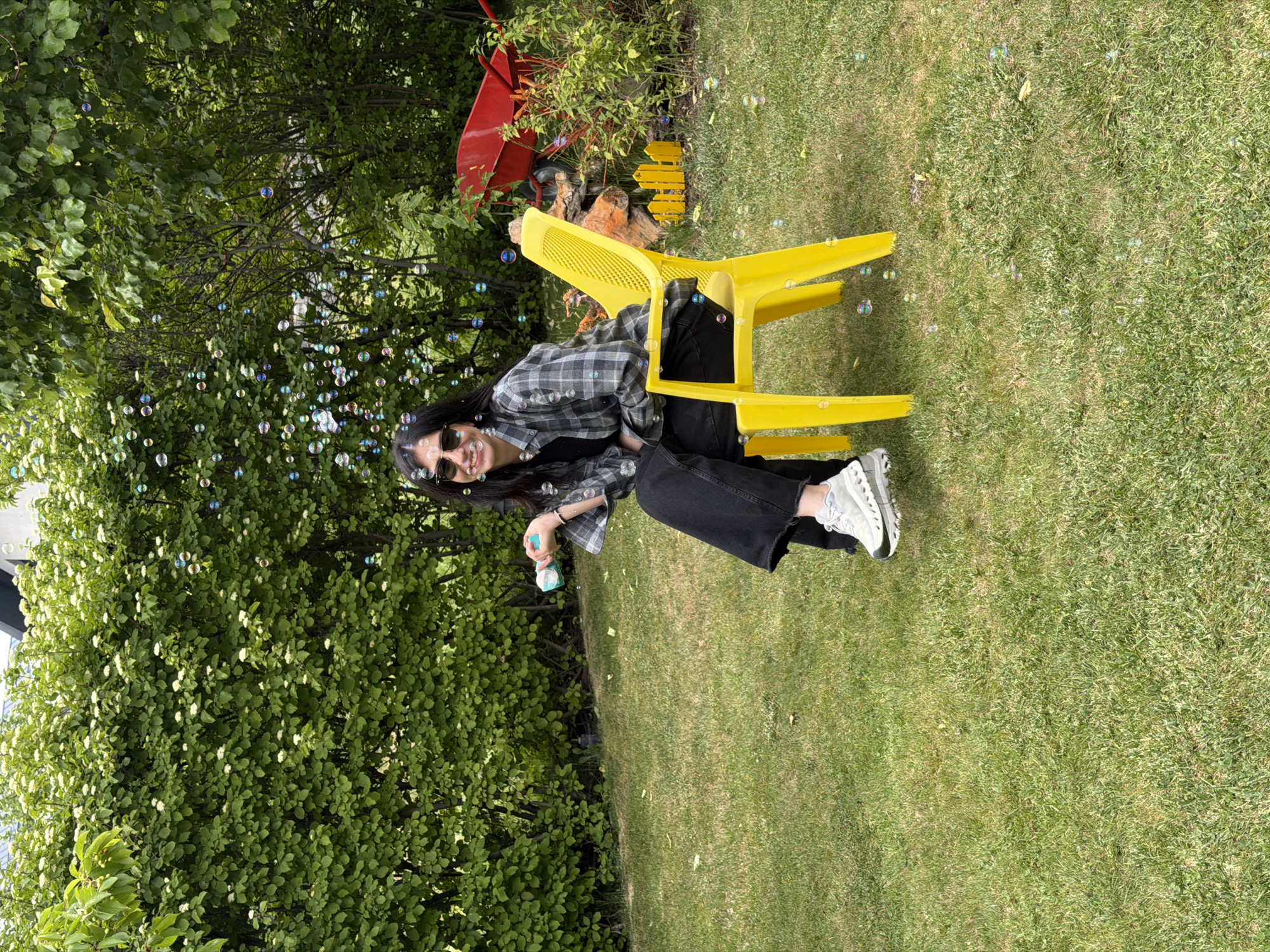 A young woman sits on a yellow plastic chair in a grassy outdoor area, possibly a yard or park. She wears a plaid shirt, dark pants, and sunglasses. She is blowing bubbles from a wand, which float around her. To her left is dense green foliage. In the background, a red wheelbarrow and a small yellow fence-like decoration are visible. The location is Yerevan, Armenia.