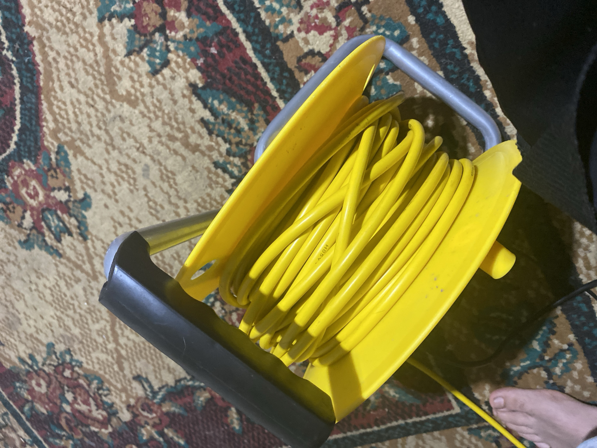 A bright yellow extension cord is coiled on a yellow reel with a black handle and gray metal frame. The reel sits on a patterned carpet with a mix of brown, red, and blue colors. In the lower left corner, a person's bare foot is visible, and a yellow cord extends from the reel towards it. The scene appears to be indoors, in an apartment or house located in Shakhtinsk, Kazakhstan.