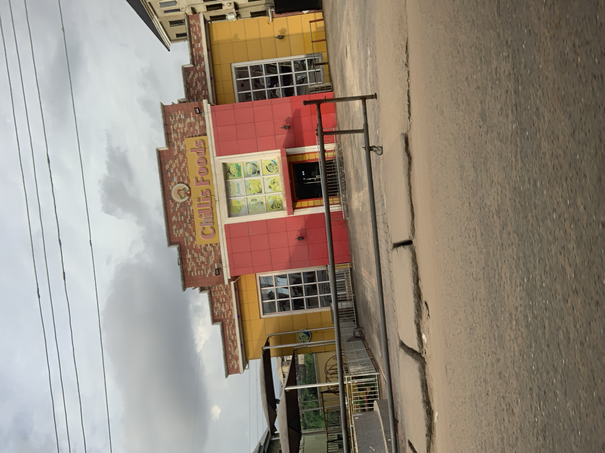 The image depicts the exterior of "Chilli's Foods" restaurant located in Nkpor, Nigeria. The building features a distinctive facade with sections painted in bright red and yellow, topped with a brick-patterned roofline. A prominent sign above the entrance reads "Chilli's Foods" in yellow lettering. Large windows are visible on both the red and yellow sections of the building. In front of the restaurant, a metal barrier with a wheel is present, and to the left, an outdoor seating area with canopies is set up. The ground in the foreground is paved and appears to be a road or sidewalk, with some cracks and debris. The sky above is overcast with scattered clouds. No people are clearly visible in the image.