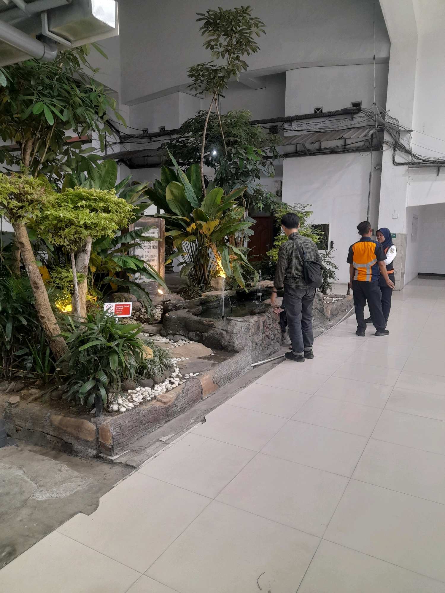 In Surabaya, Indonesia, an indoor oasis features a man in a checkered shirt and dark pants, carrying a black satchel, interacting with a small decorative pond. Two individuals in uniform, one with an orange vest and the other in a blue shirt and headscarf, stand nearby, appearing to converse. Lush green plants and trees surround the pond, creating a naturalistic display. White tiles line the floor, and conduits and lighting fixtures are visible on the ceiling, indicating an interior setting, possibly a public building or office space.