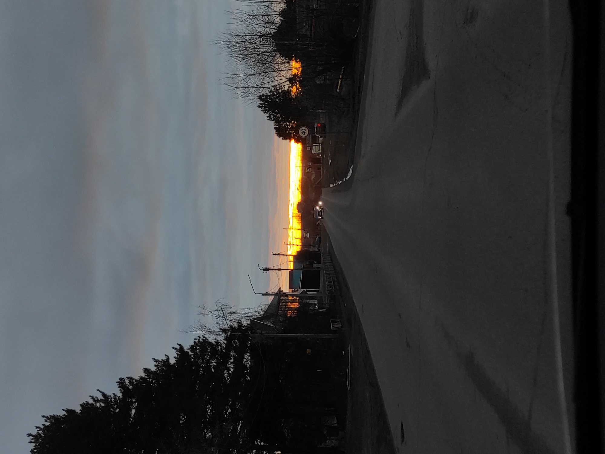 The image depicts a straight road at what appears to be sunset or sunrise in Chaikovsky, Russia. The sky above is a mix of muted blue and grey clouds, but a striking, vibrant horizontal strip of intense orange and yellow light dominates the distant horizon, indicating the sun breaking through.
The asphalt road extends into the distance, leading towards this brilliant glow. Along both sides of the road, dark silhouettes of numerous trees are visible, some with bare, intricate branches and others with denser foliage. On the left side of the road, a street light pole stands tall. Further down, buildings or structures are partially visible as dark shapes, catching faint reflections of the bright sky.
A clear circular speed limit sign, featuring the number "40" in black on a white background with a red border, is positioned on the left. Below it, a triangular warning sign with a red border is also visible, though its specific symbol is indistinct. In the mid-distance, a car with illuminated headlights is seen driving away from the viewer, heading towards the radiant horizon. The scene conveys a sense of quietude, with the dramatic natural light contrasting against the shadowy foreground elements.